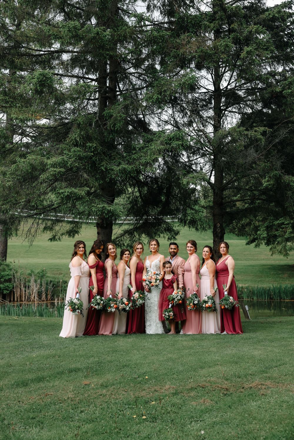 Wedding party in pink and burgundy dresses pose together under evergreen trees.