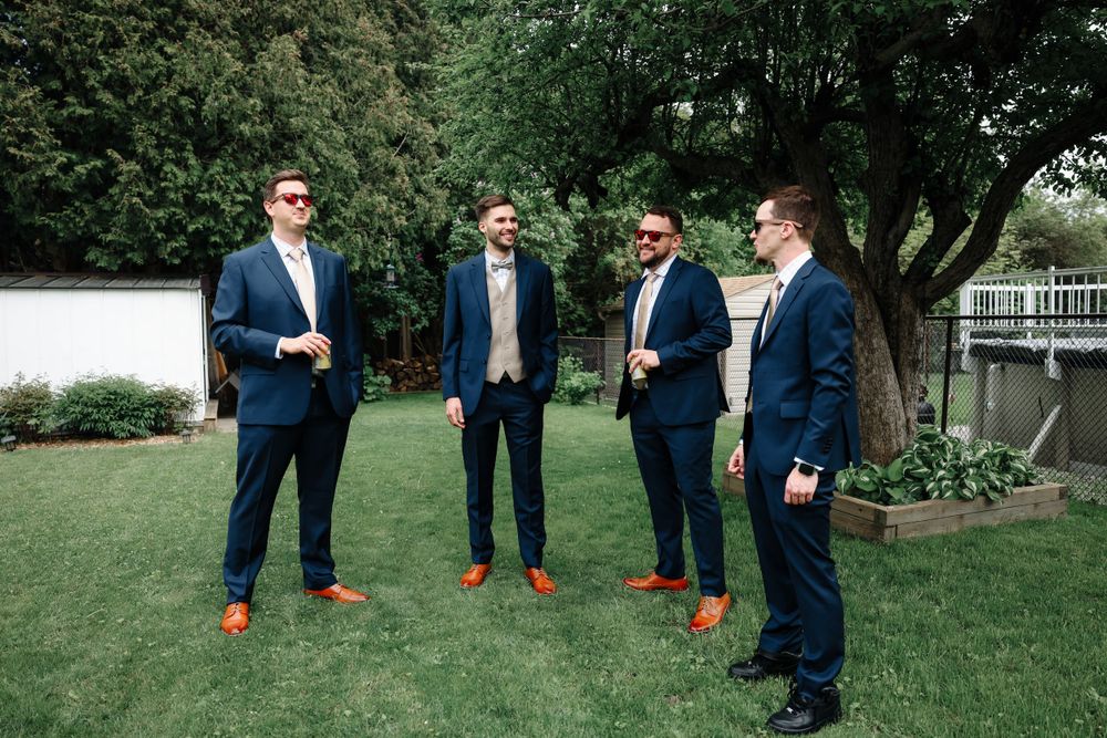 Groomsmen in matching navy suits with brown shoes standing on manicured lawn.