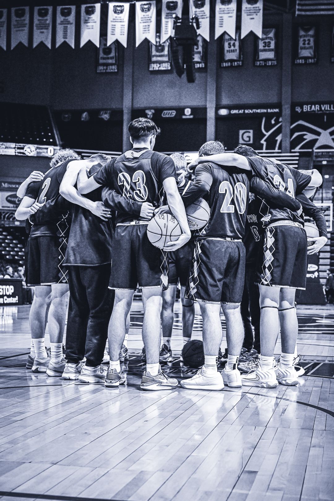 Basketball team forms huddle on court with championship banners visible in background.