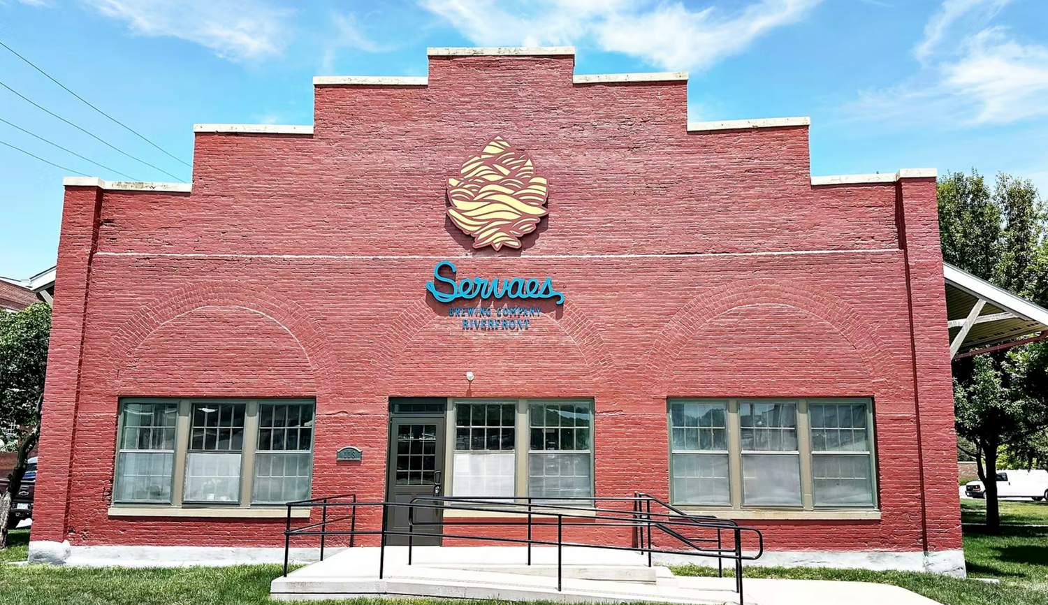 A brick building with a stepped roofline, featuring a large emblem and the word Servaes with brewing below in blue letters.
