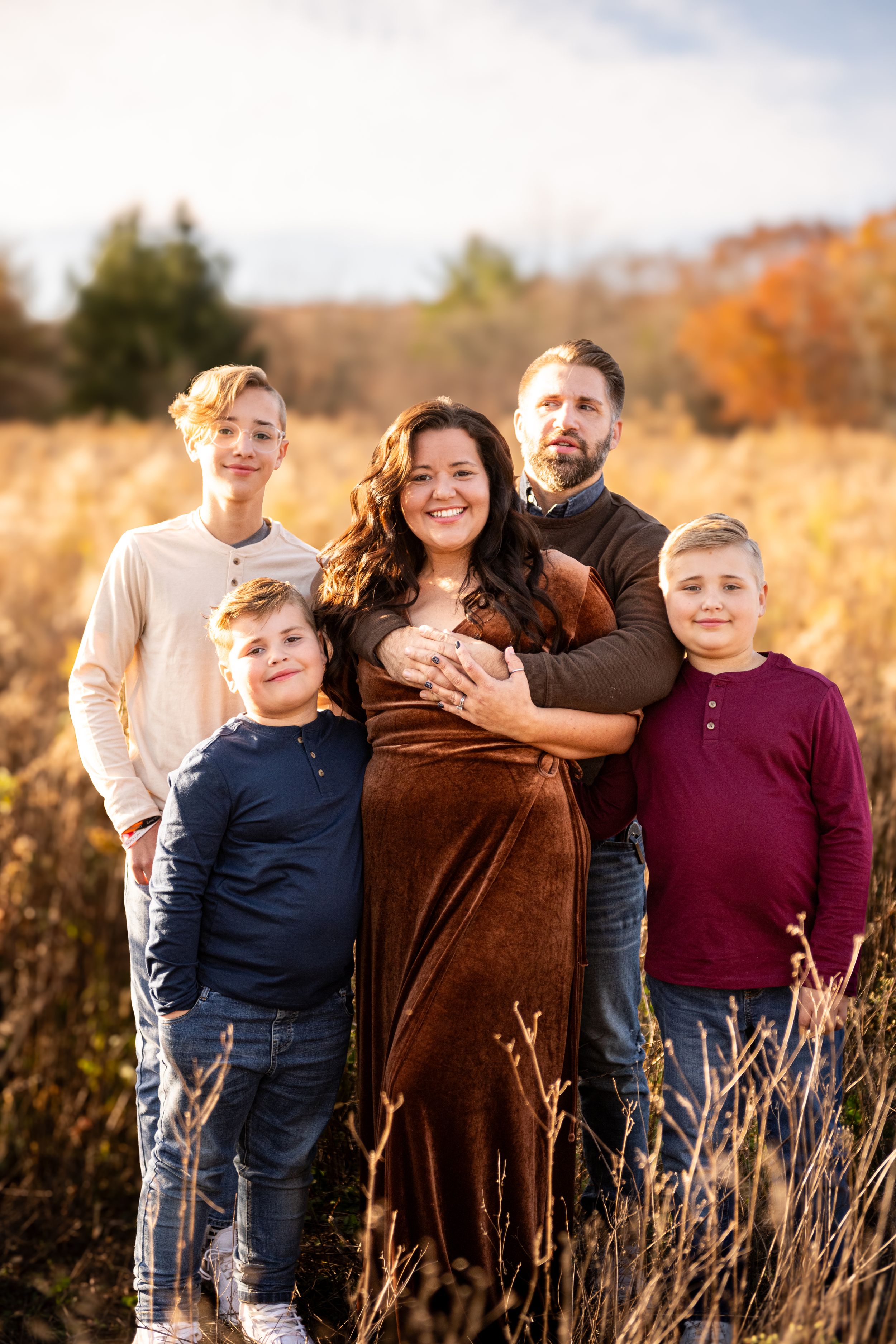 Family of five poses together in a golden autumn field during sunset wearing casual clothing.