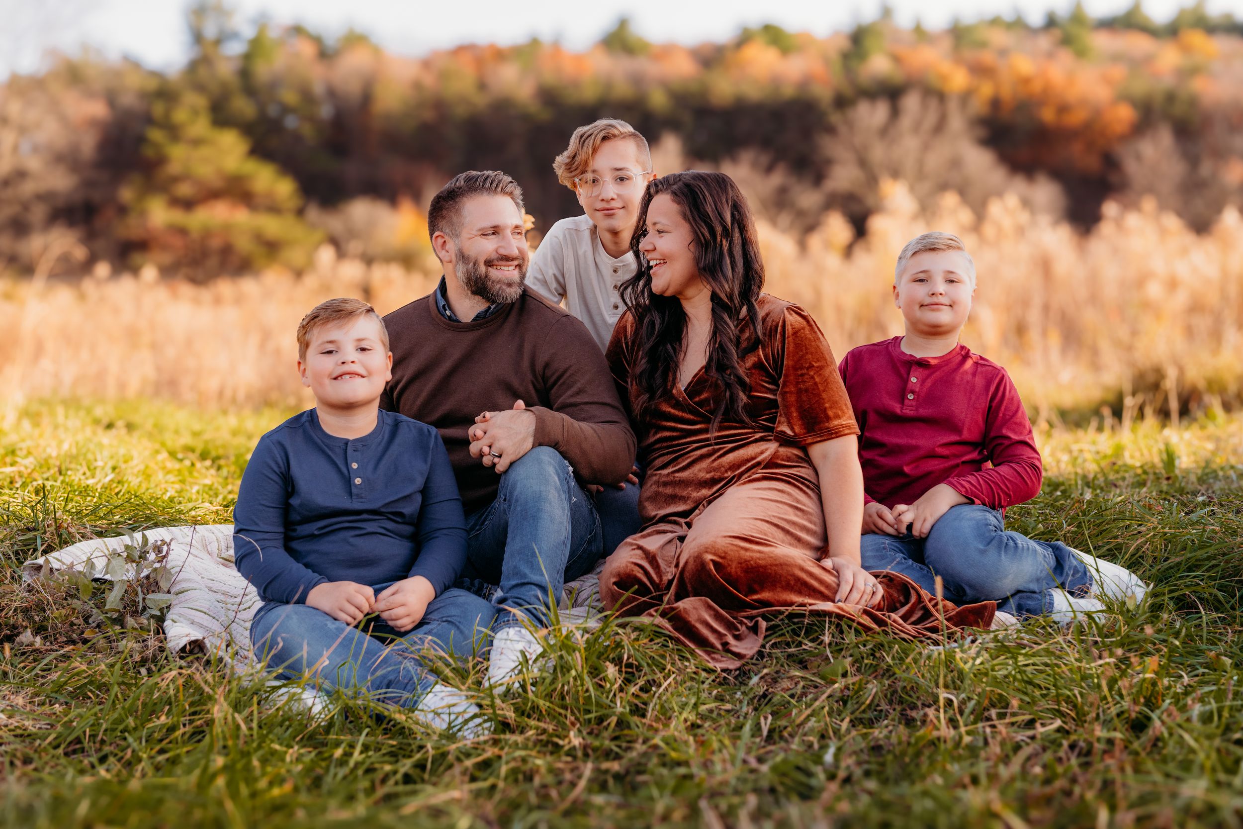 A family sits together on grassy hill during golden hour with autumn foliage in background.