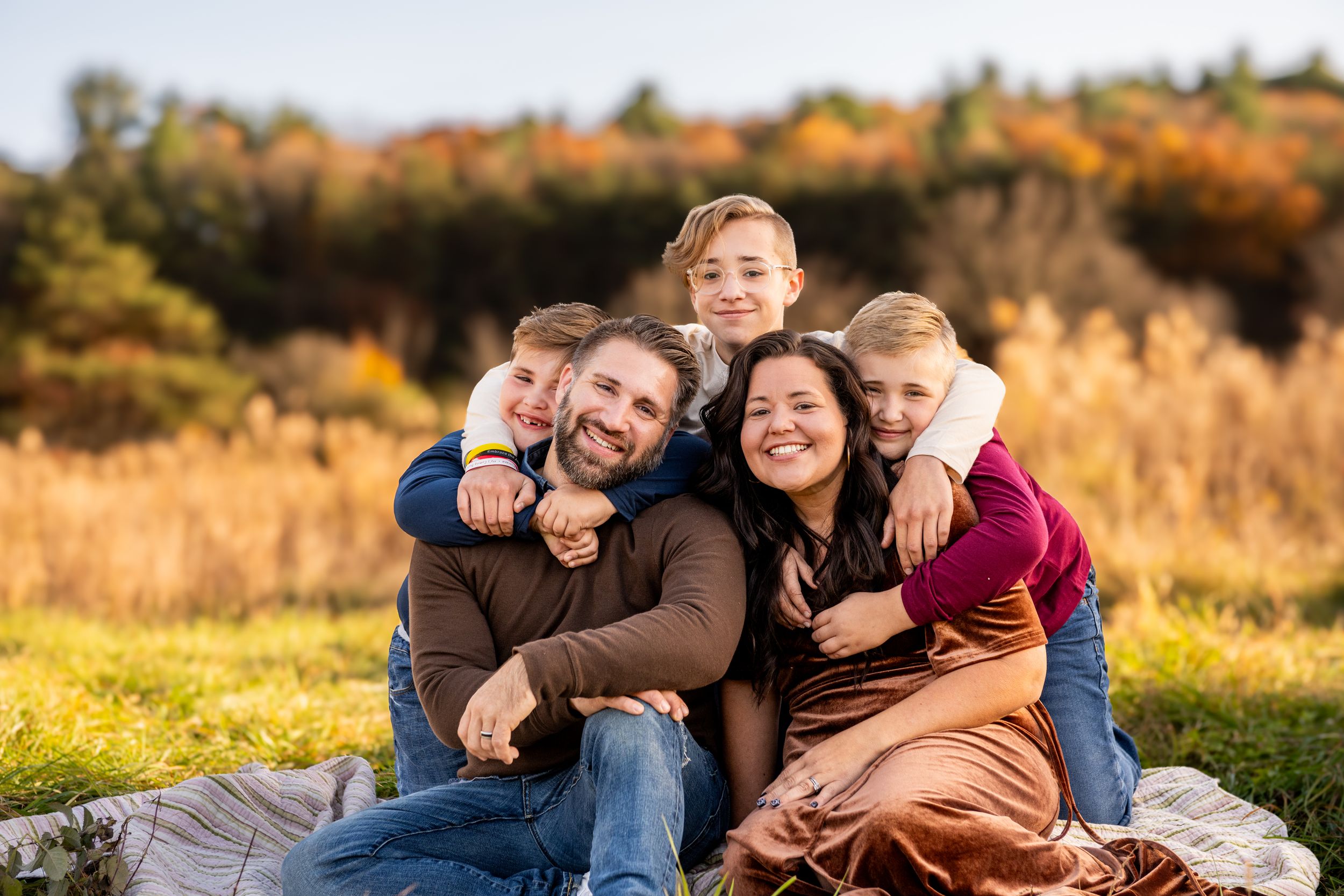 Happy family group embraces while sitting on blanket in autumn field with golden grass and foliage.