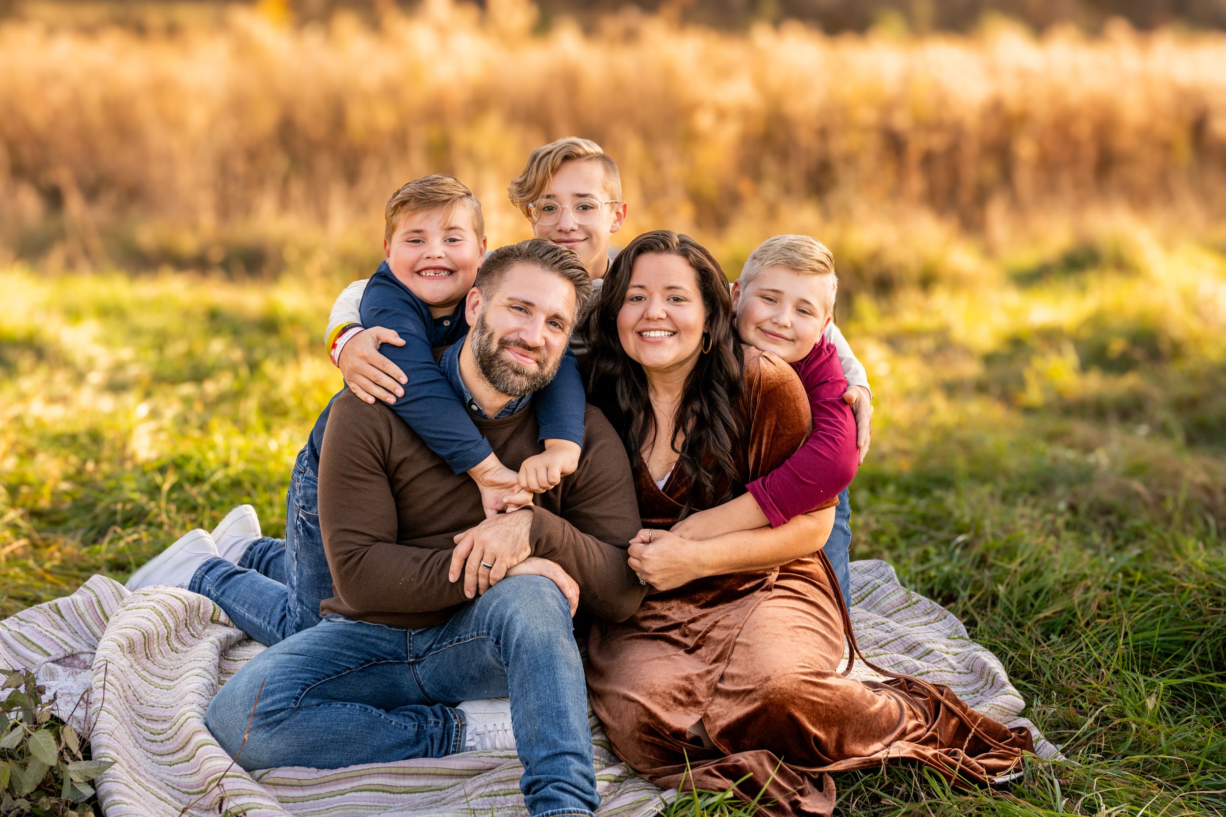 A loving family sits together on a blanket in a grassy meadow during golden hour sunset.