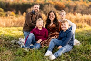 A family of five sits together on grass during golden hour in an autumn field.