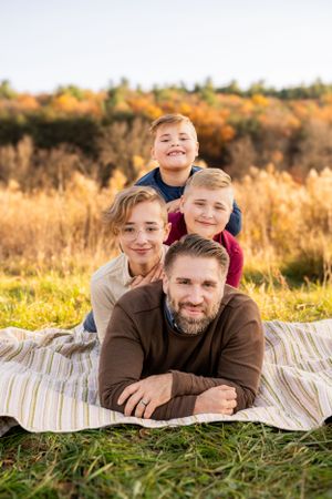 A family lies together on a striped blanket in a sunny autumn field with golden grass and fall foliage in the background.