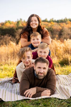 A family group poses together on a blanket outdoors in a sunlit autumn field with golden grass and trees in background.
