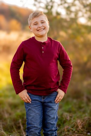 Young person wearing a burgundy henley shirt and jeans stands outdoors in warm sunlight during golden hour.