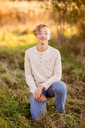 A young person in a beige henley shirt and jeans poses outdoors during golden hour sunset lighting.