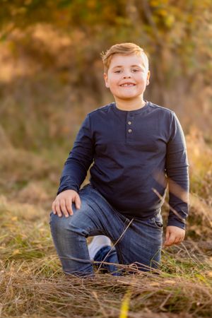A young boy in a navy henley shirt kneels in tall grass during golden hour.