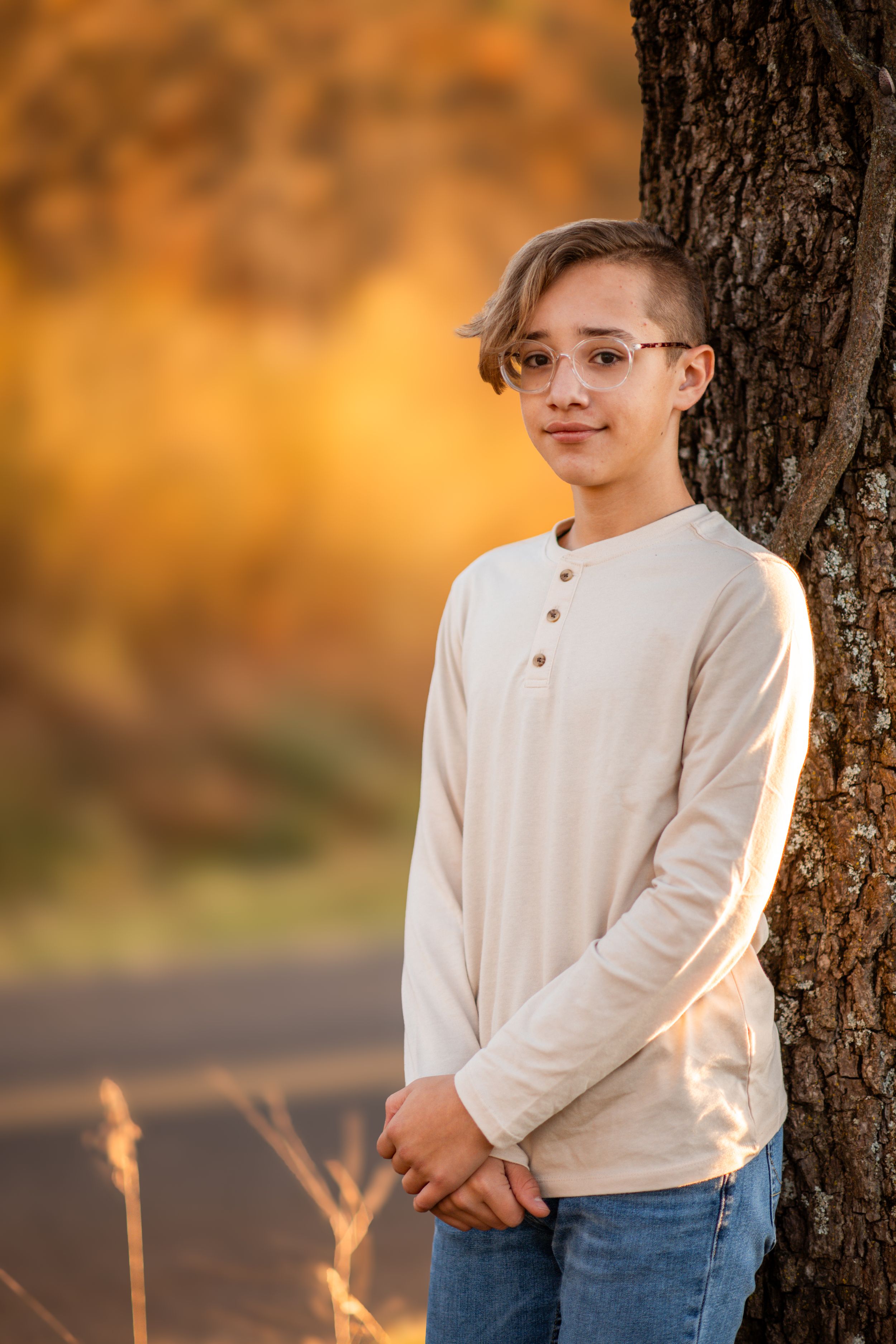 Youth in glasses and cream henley sweater poses by tree trunk against warm autumn sunset backdrop.