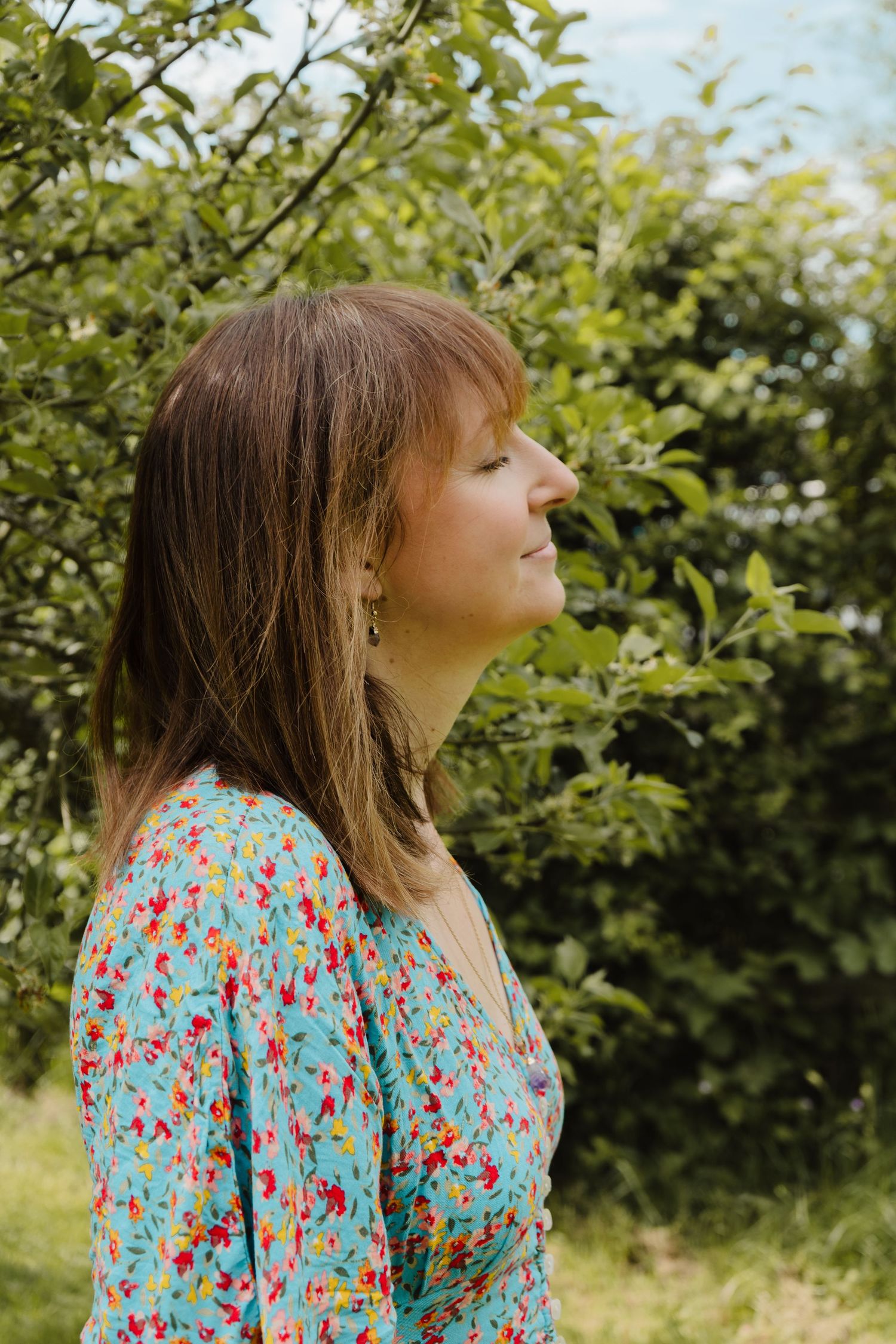 photo of a white female with brown hair amongst trees in a floral dress, smiling in a storytelling brand photoshoot for a reiki practitioner