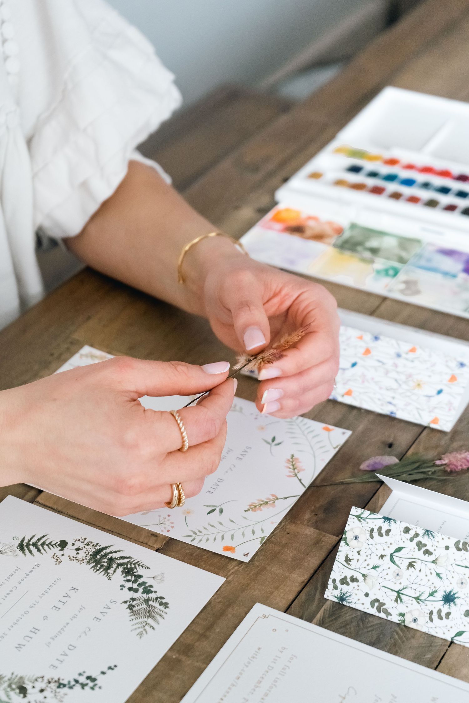 photo of an artists hands holding dried flowers, surrounded by her paints at a table