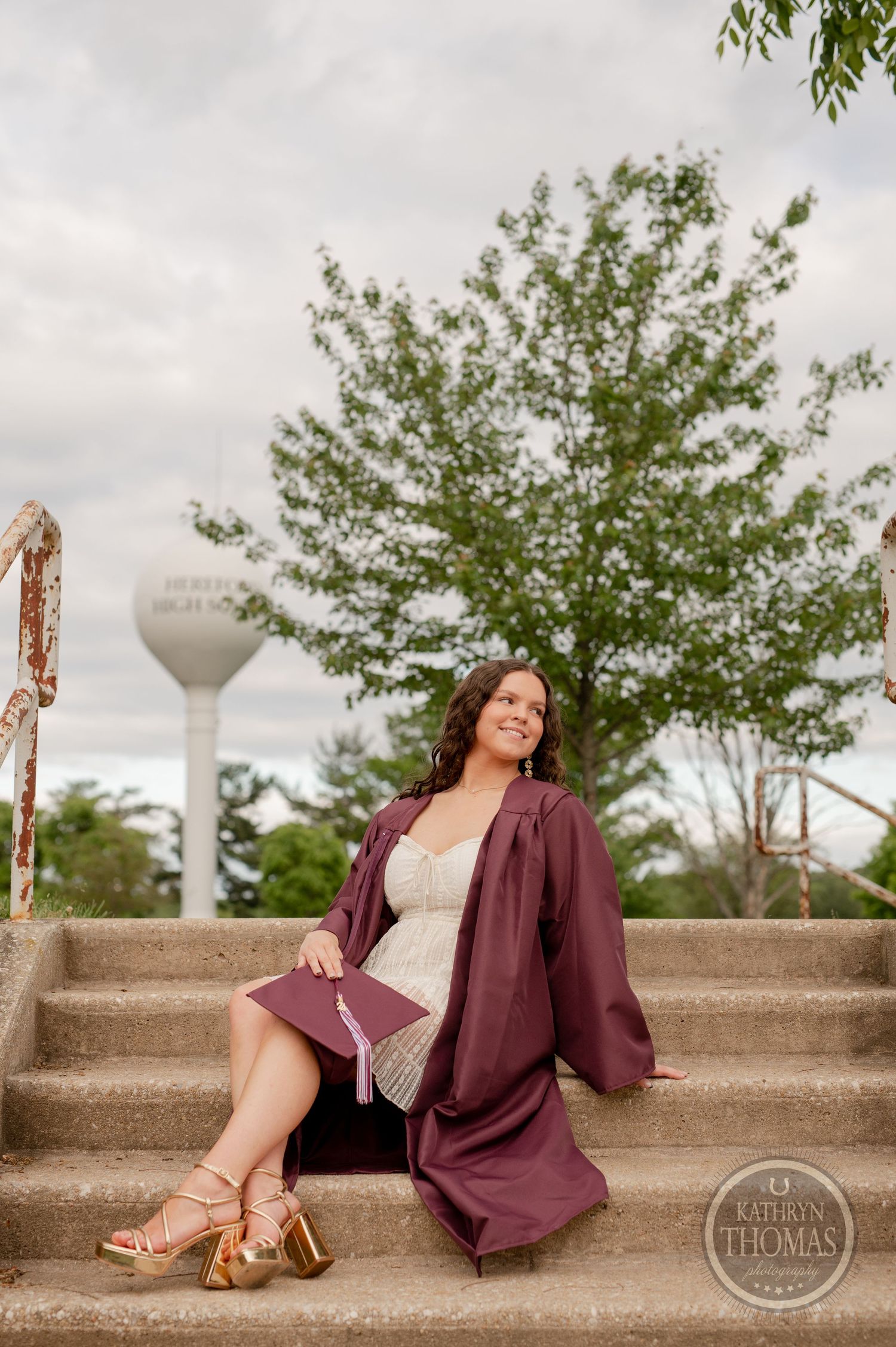 Graduate in maroon gown sits on concrete steps with water tower in background.