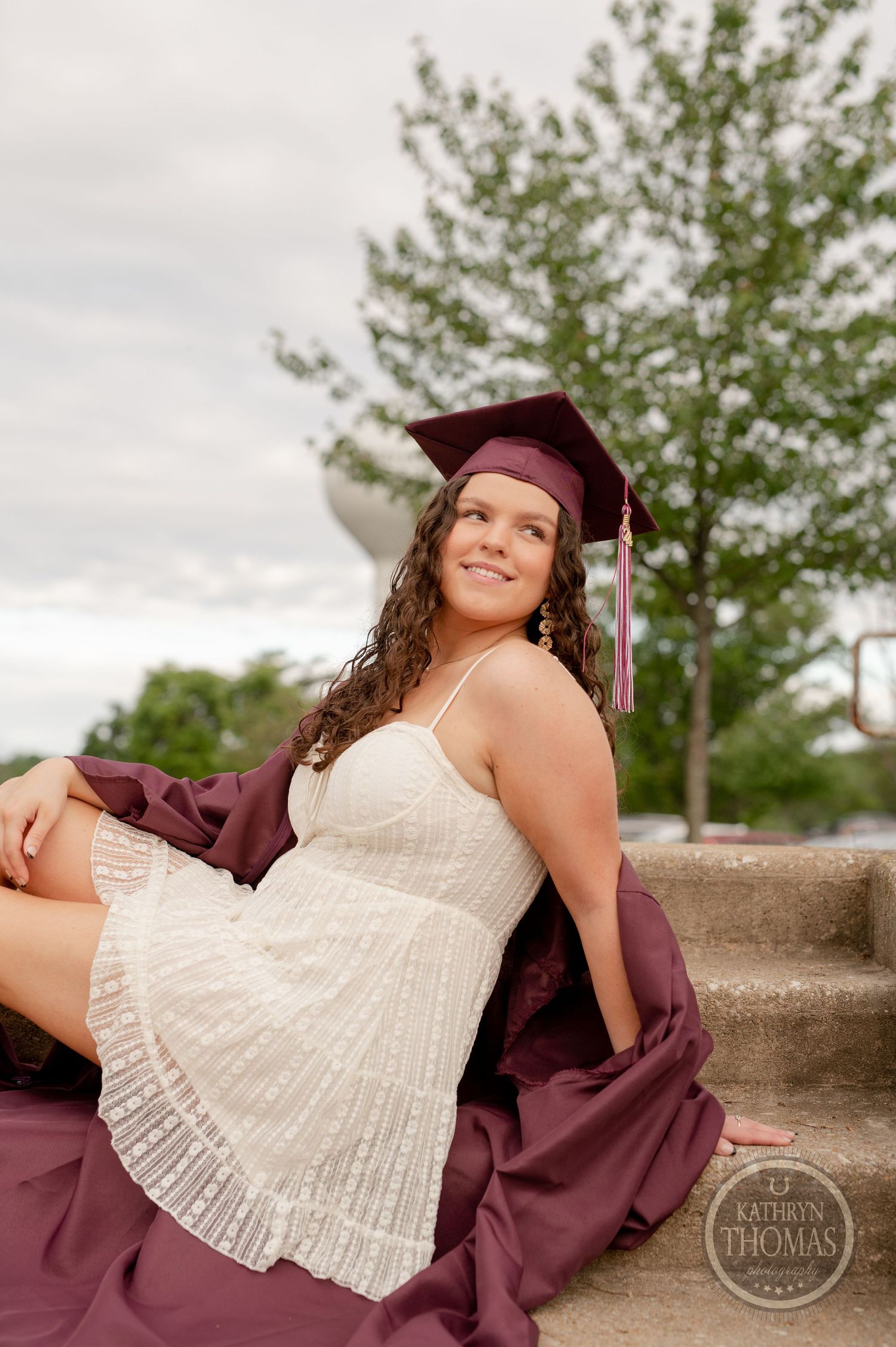 Graduate in white dress and maroon cap reclines on concrete steps outdoors.