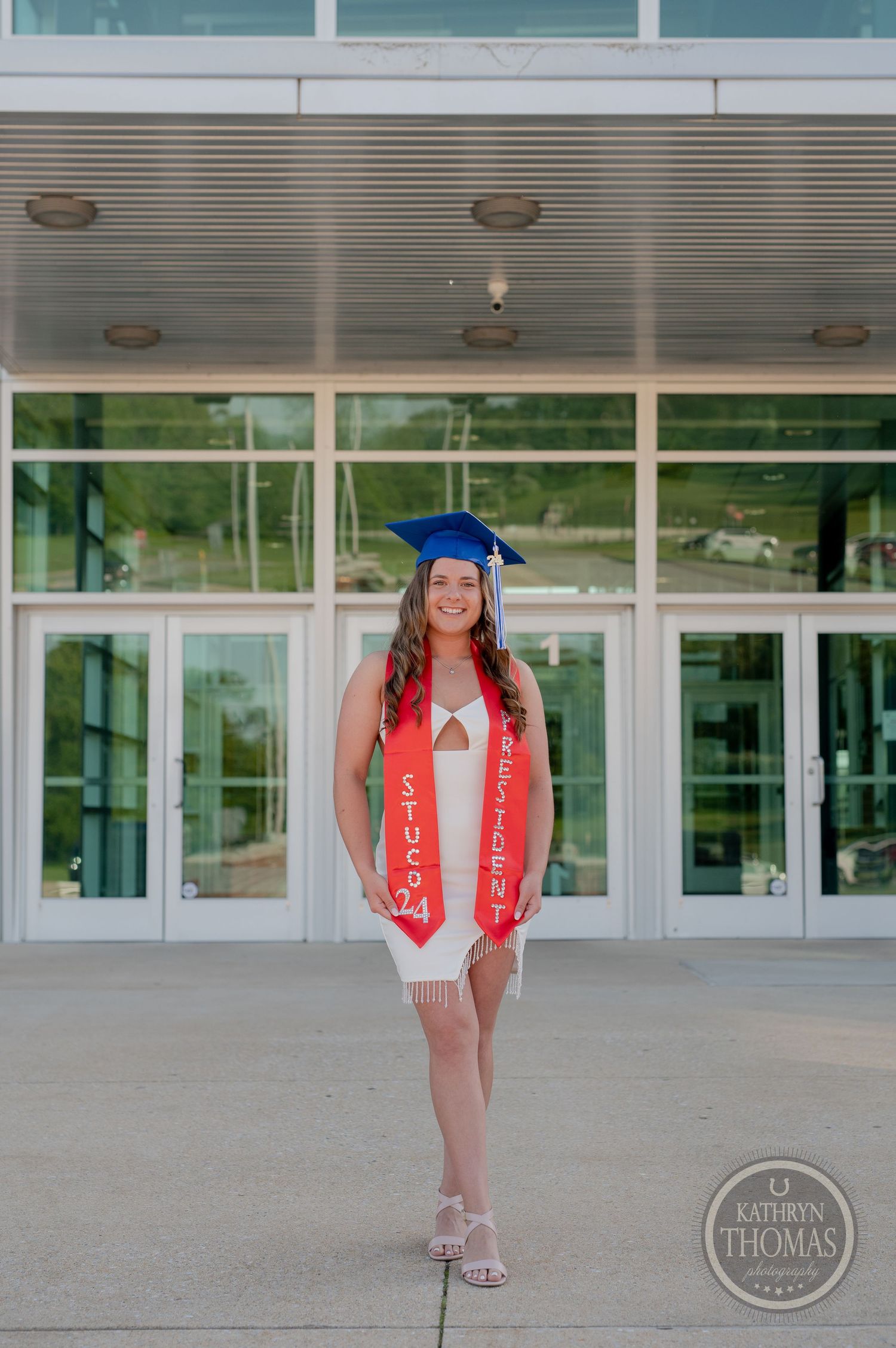 Graduate in blue cap and white dress poses in front of glass building entrance.