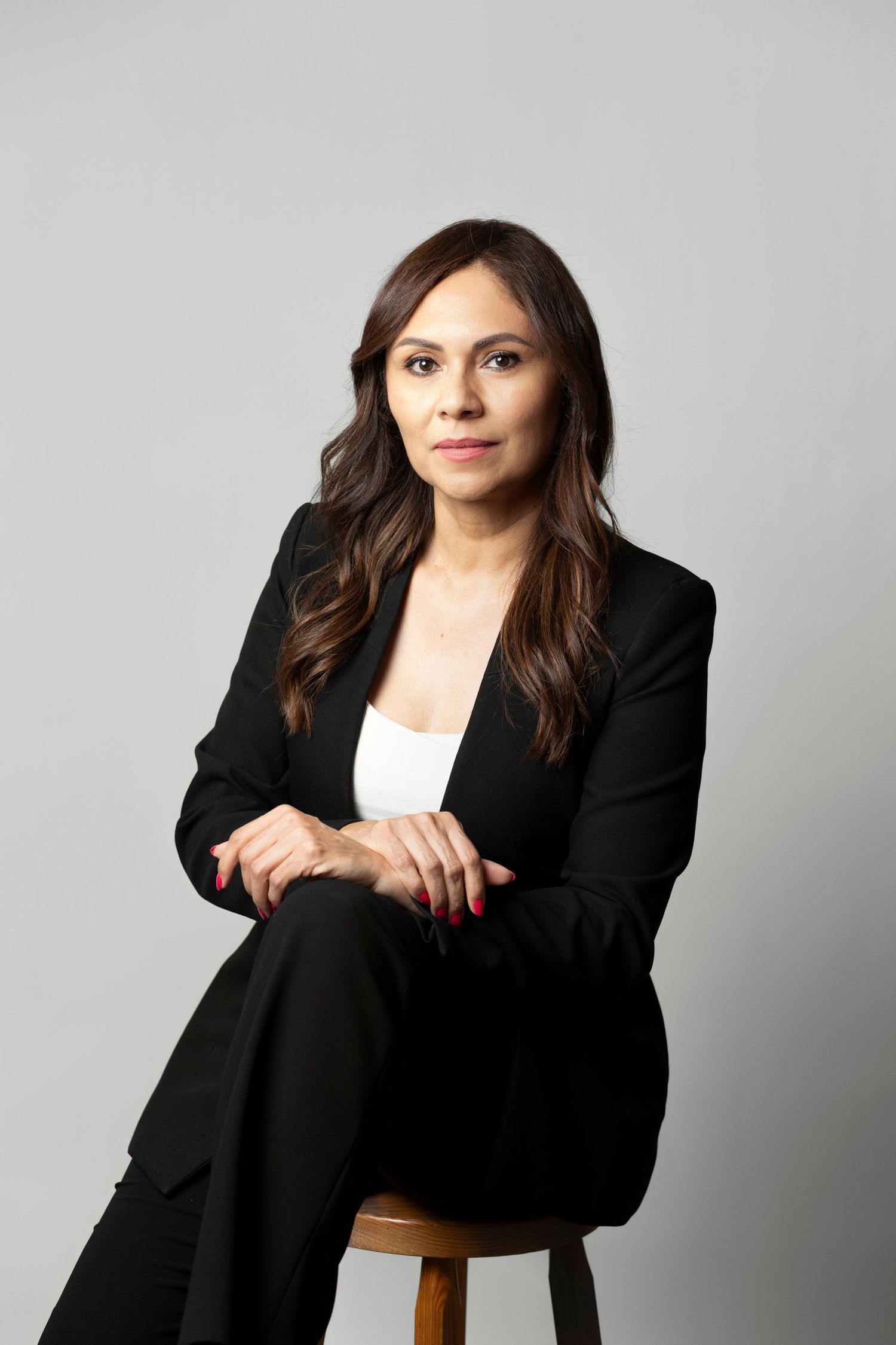 A professional woman in a black blazer sits on a stool, posing confidently against a gray background.