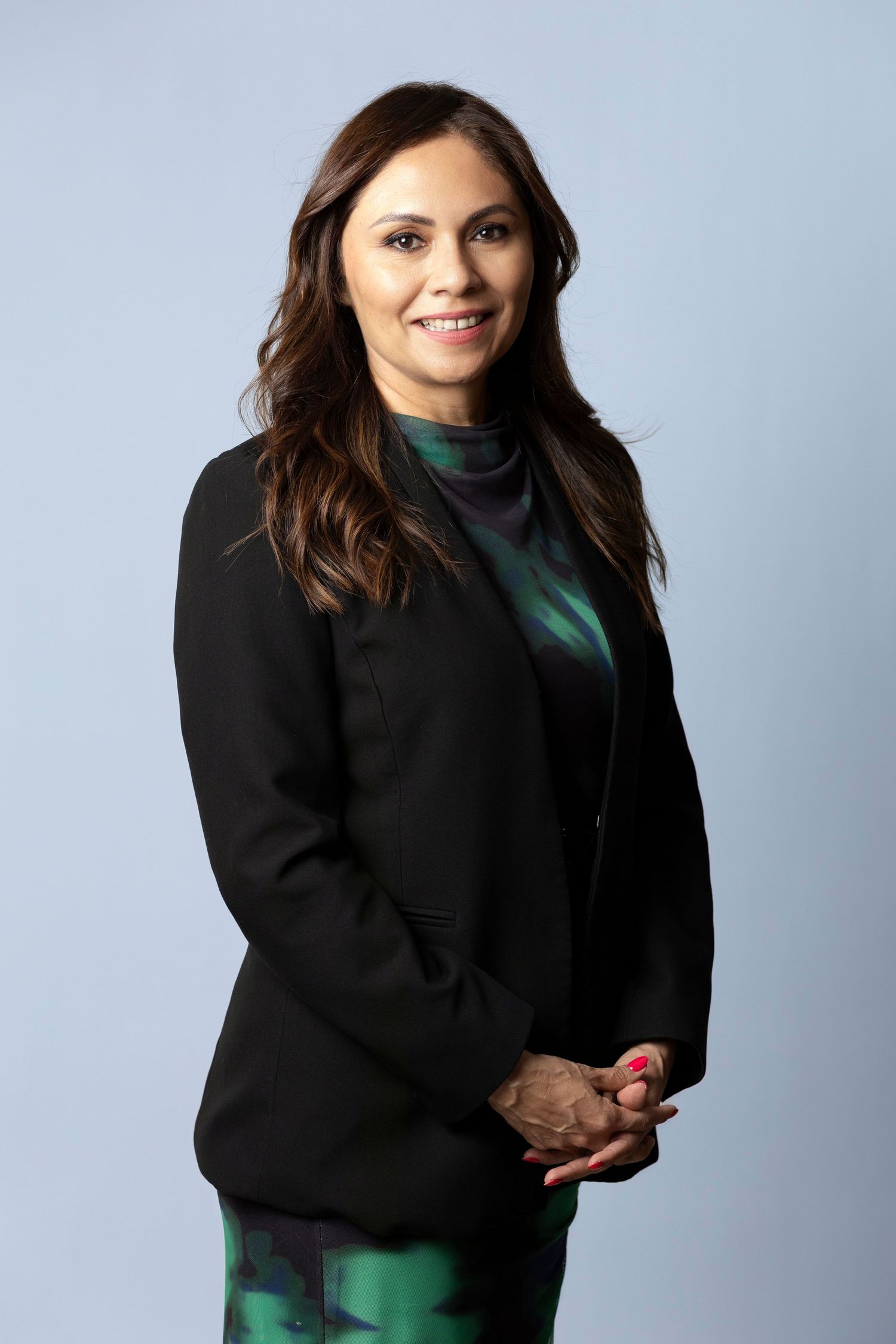 Professional woman in black blazer and green patterned dress smiling confidently against light blue background.