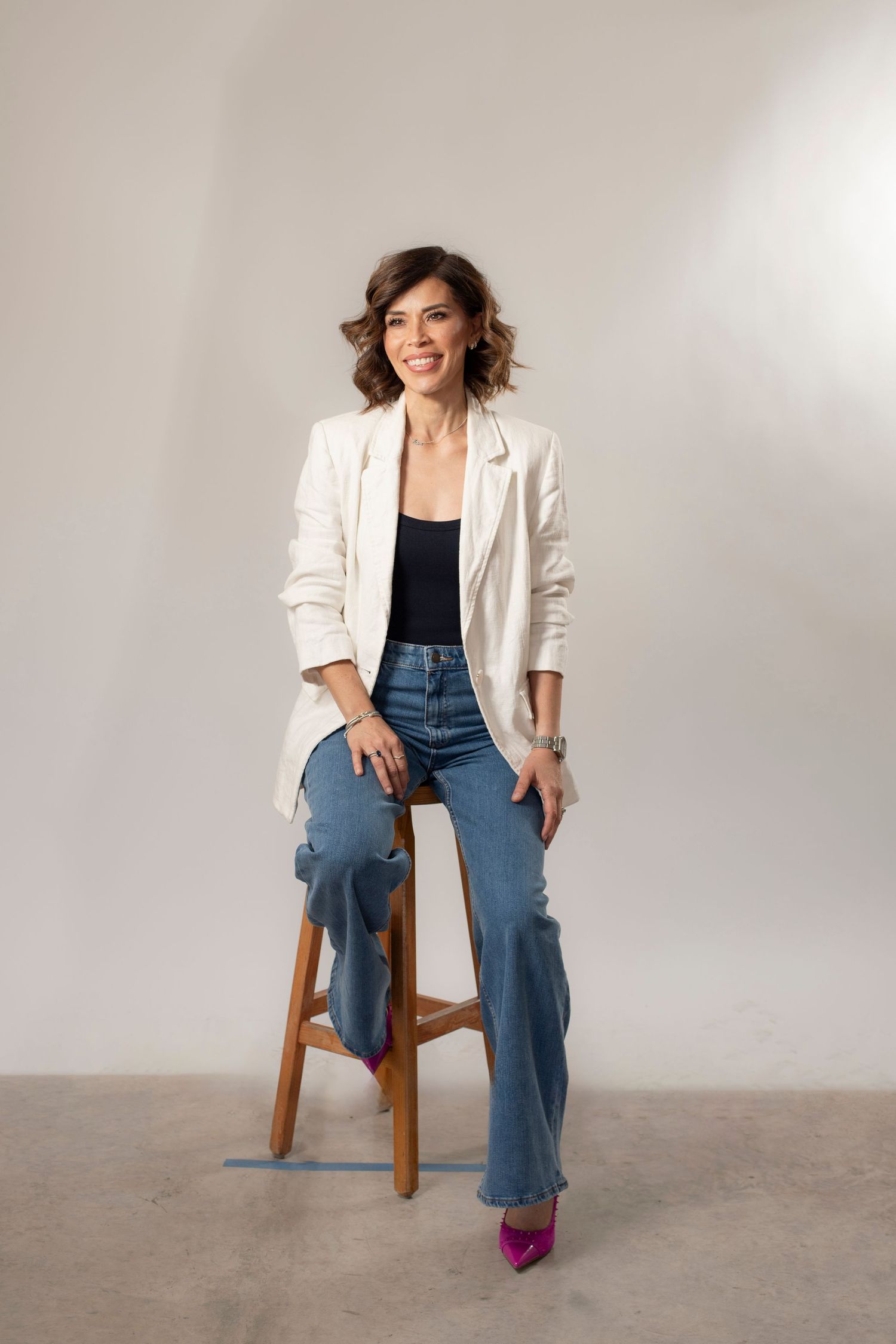 Smiling woman in white blazer, black top, jeans, and pink heels sitting on a wooden stool against a gray background.