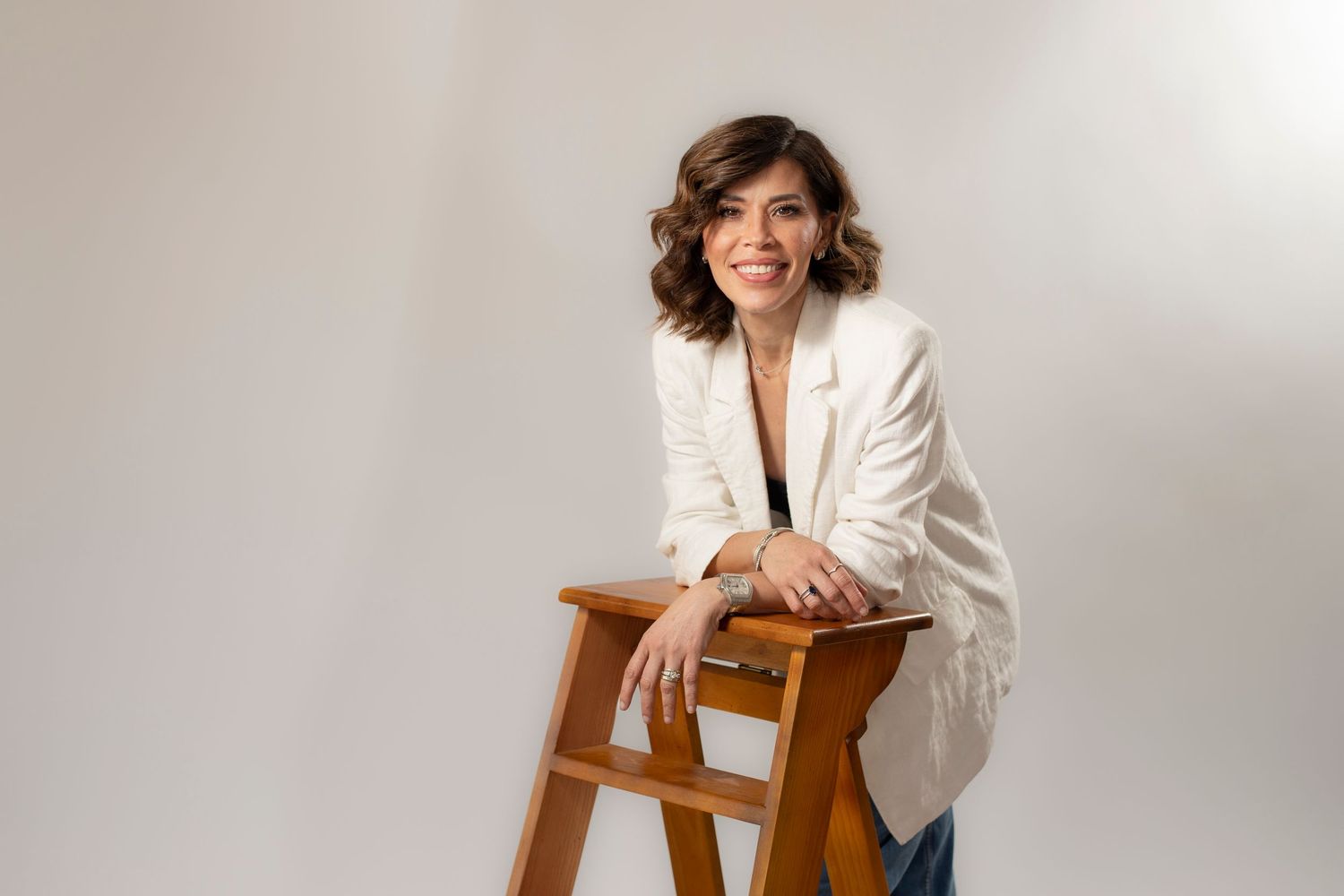 Woman in white blazer smiling, leaning on wooden stool against light gray background.