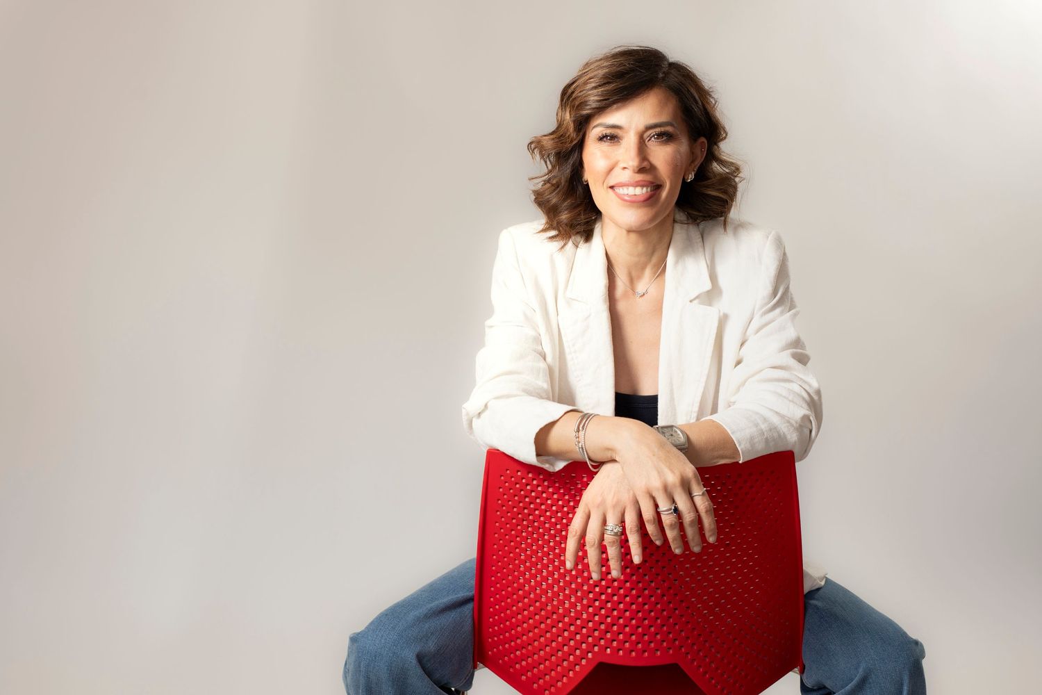 Woman in white blazer smiling, seated on red chair against light gray background.