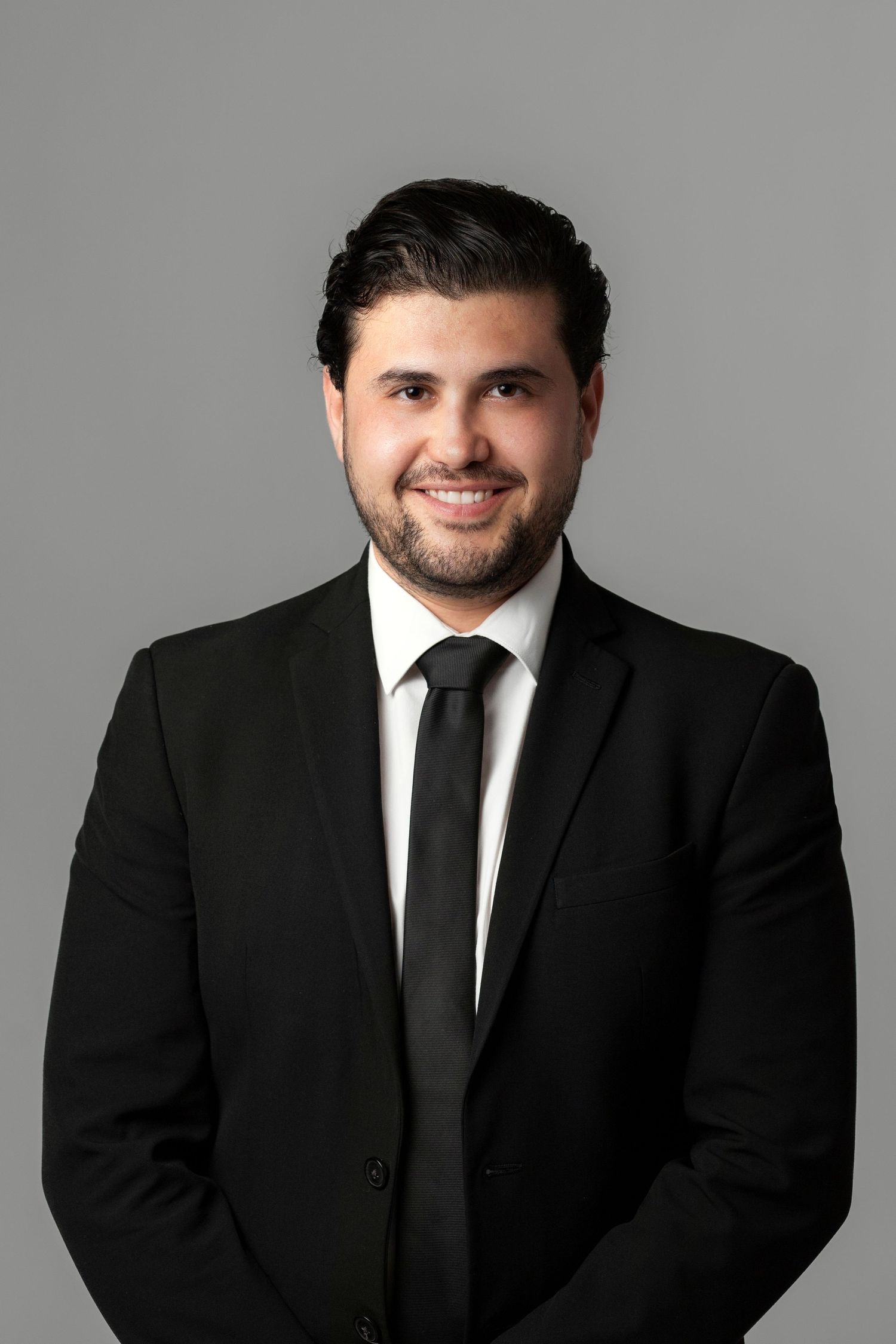 Professional headshot of a smiling young man with dark hair and beard wearing a black suit and tie.