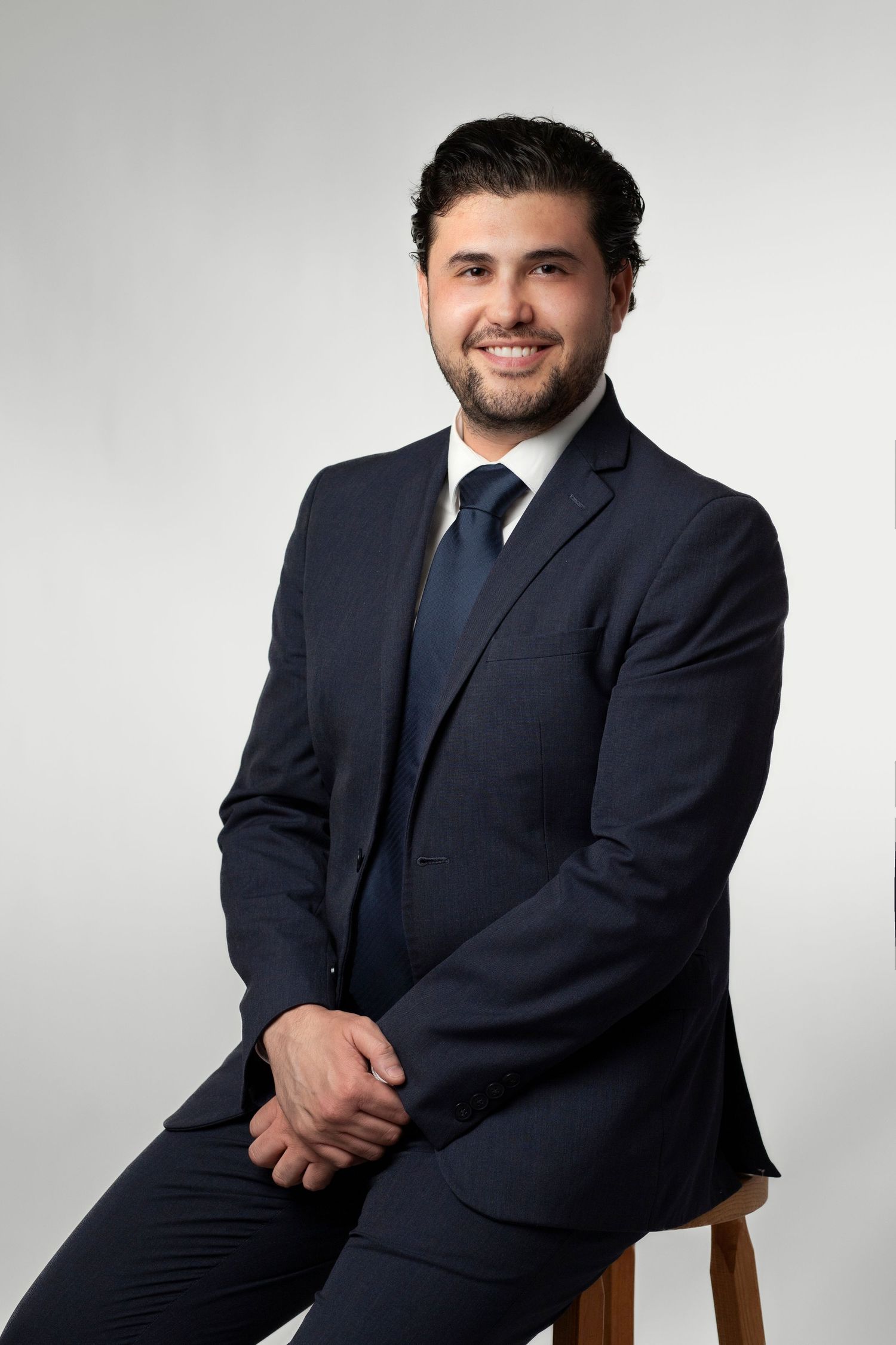 Professional man in navy suit and tie sitting confidently on stool, smiling, against light gray background.