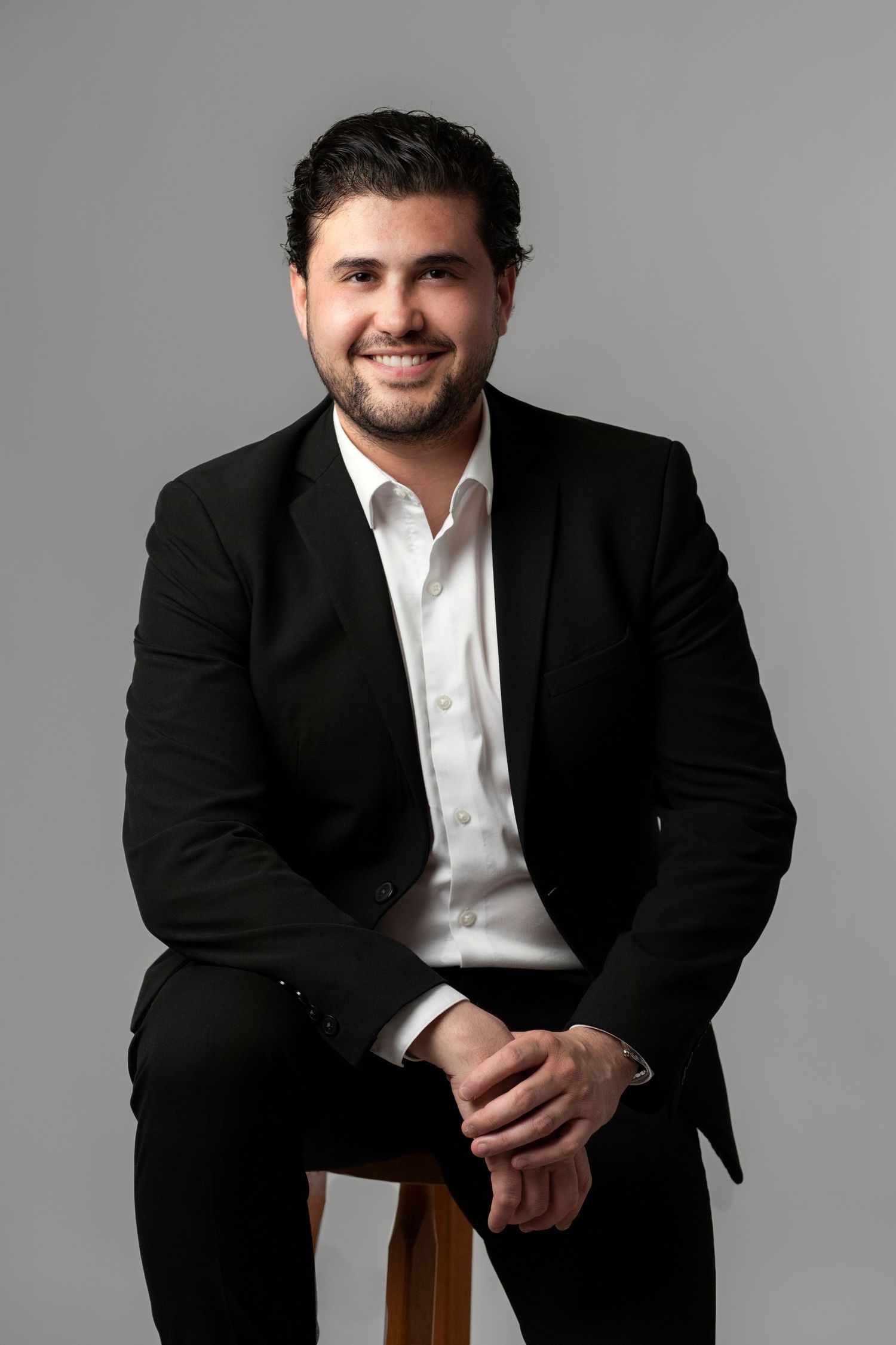 A smiling man in a black suit and white shirt sits on a stool against a gray background.