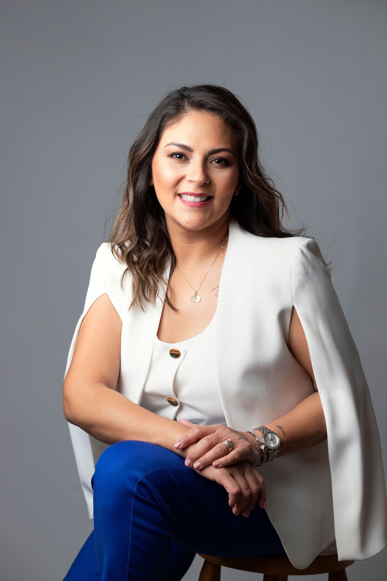 Professional woman in white blazer and blue pants sitting confidently on a stool against a gray background.