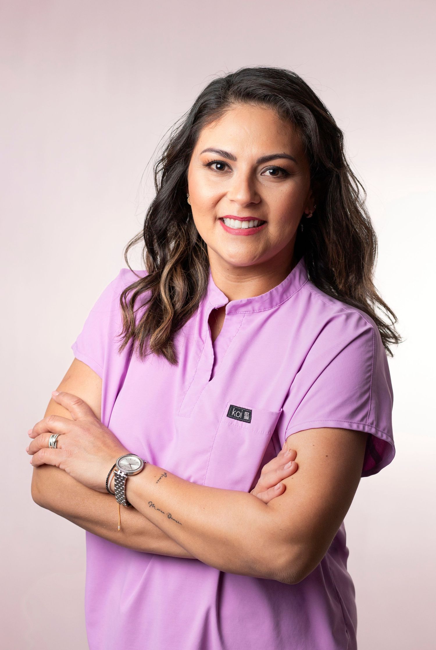Smiling female healthcare professional in lavender scrubs with arms crossed against a pink background.