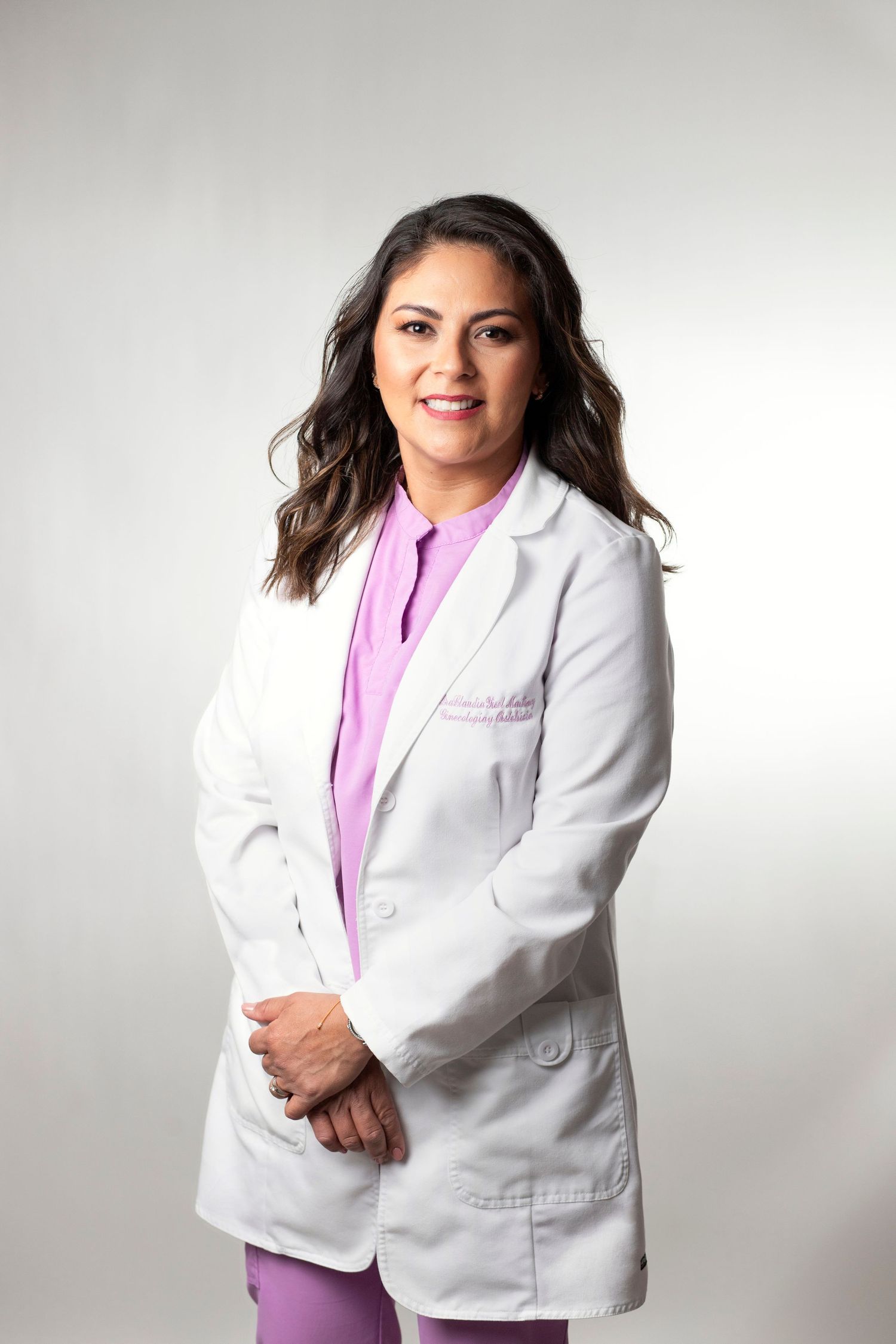 Female healthcare professional in white lab coat and pink scrubs smiling confidently in a studio portrait.