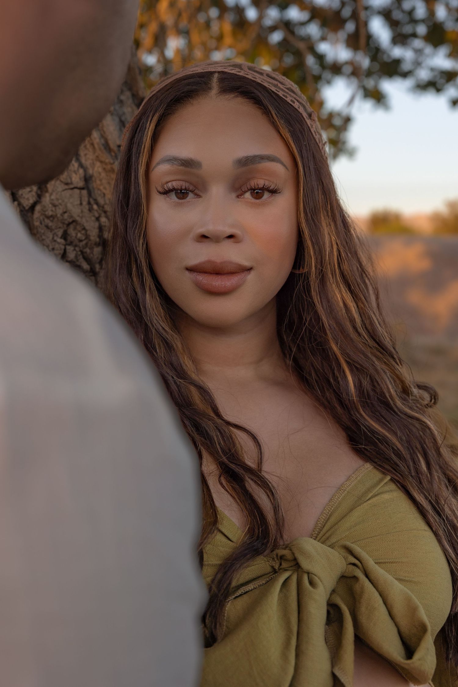 Portrait shot in golden hour lighting featuring a soft smile and long dark hair, wearing an olive green off-shoulder top.