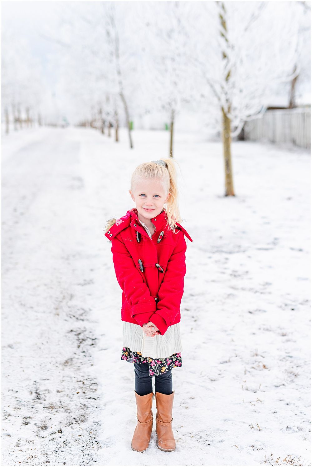 Child in red winter coat and brown boots standing on snowy path among bare trees.