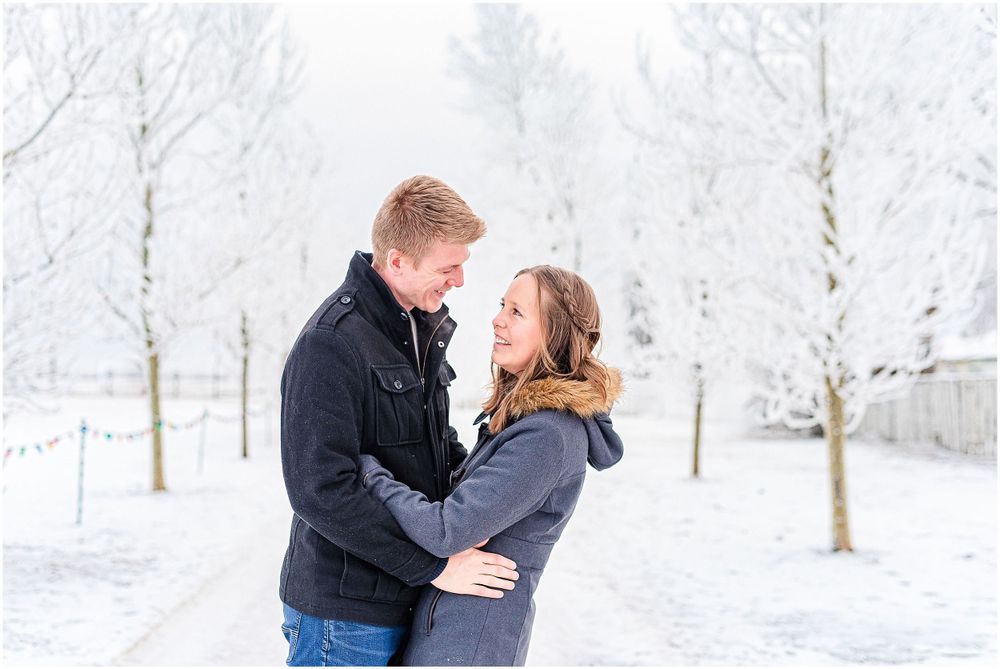 Couple embracing on snow-covered path surrounded by frost-covered trees.