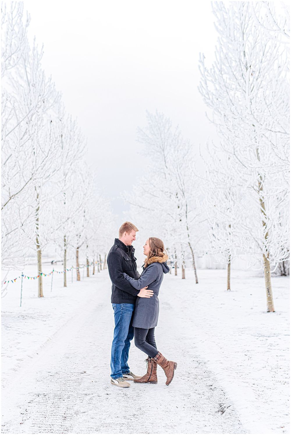 Couple embracing on snowy path surrounded by frost-covered trees in winter wonderland setting.