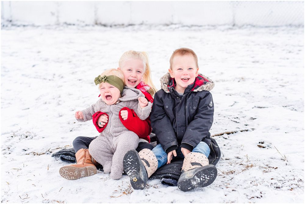 Children sitting together and laughing in the snow wearing winter coats.