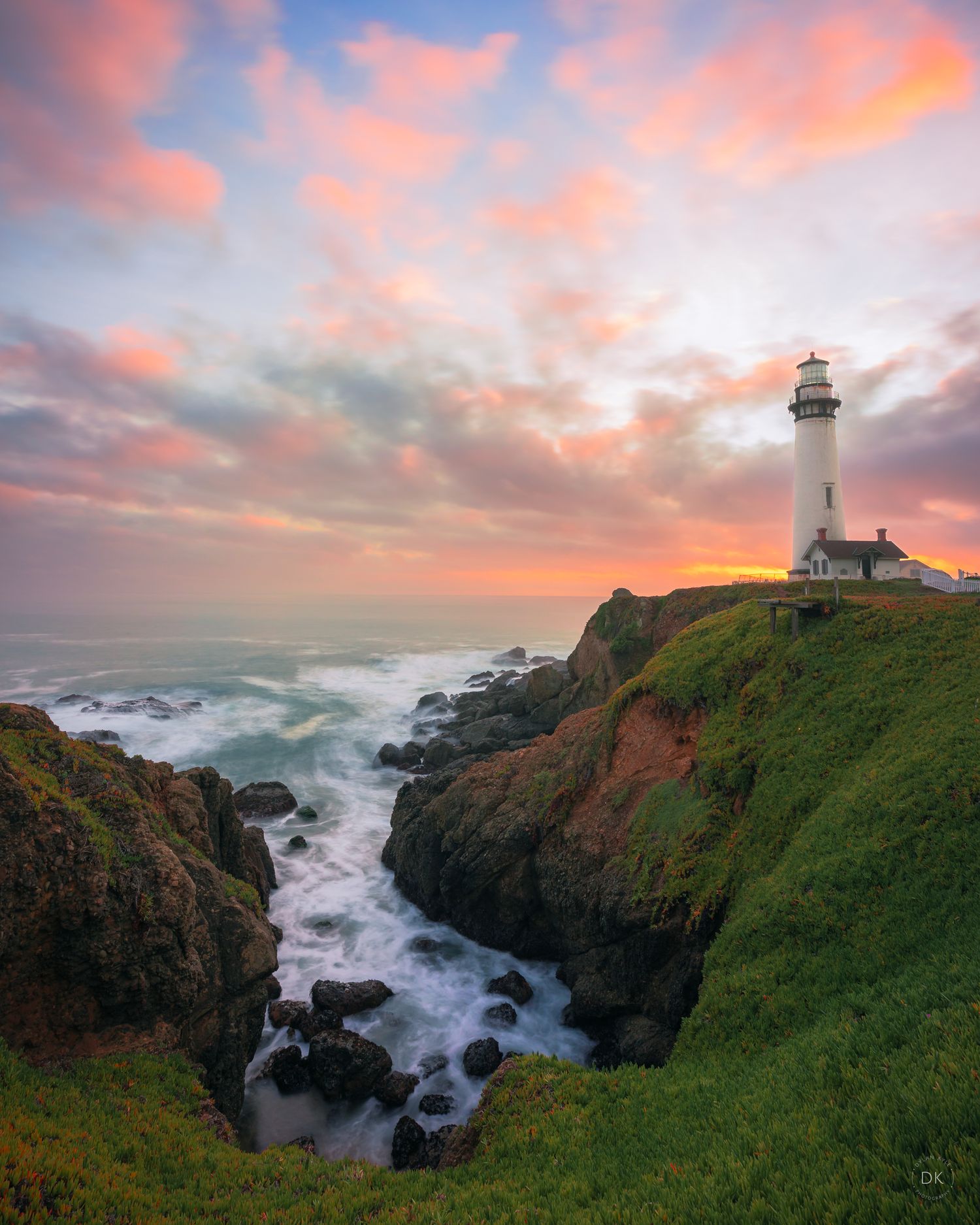 Lighthouse perched on dramatic coastal cliffs with crashing waves and vibrant pink sunset clouds over the Pacific Ocean.