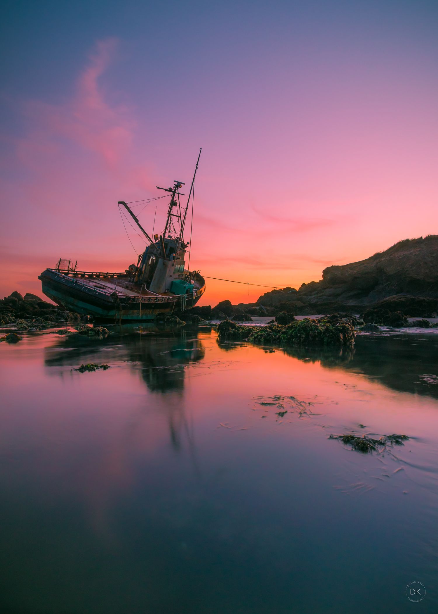 An abandoned fishing boat rests on rocks during a stunning pink and purple sunset with reflections in the calm water. Cayucos Boat Wreck.