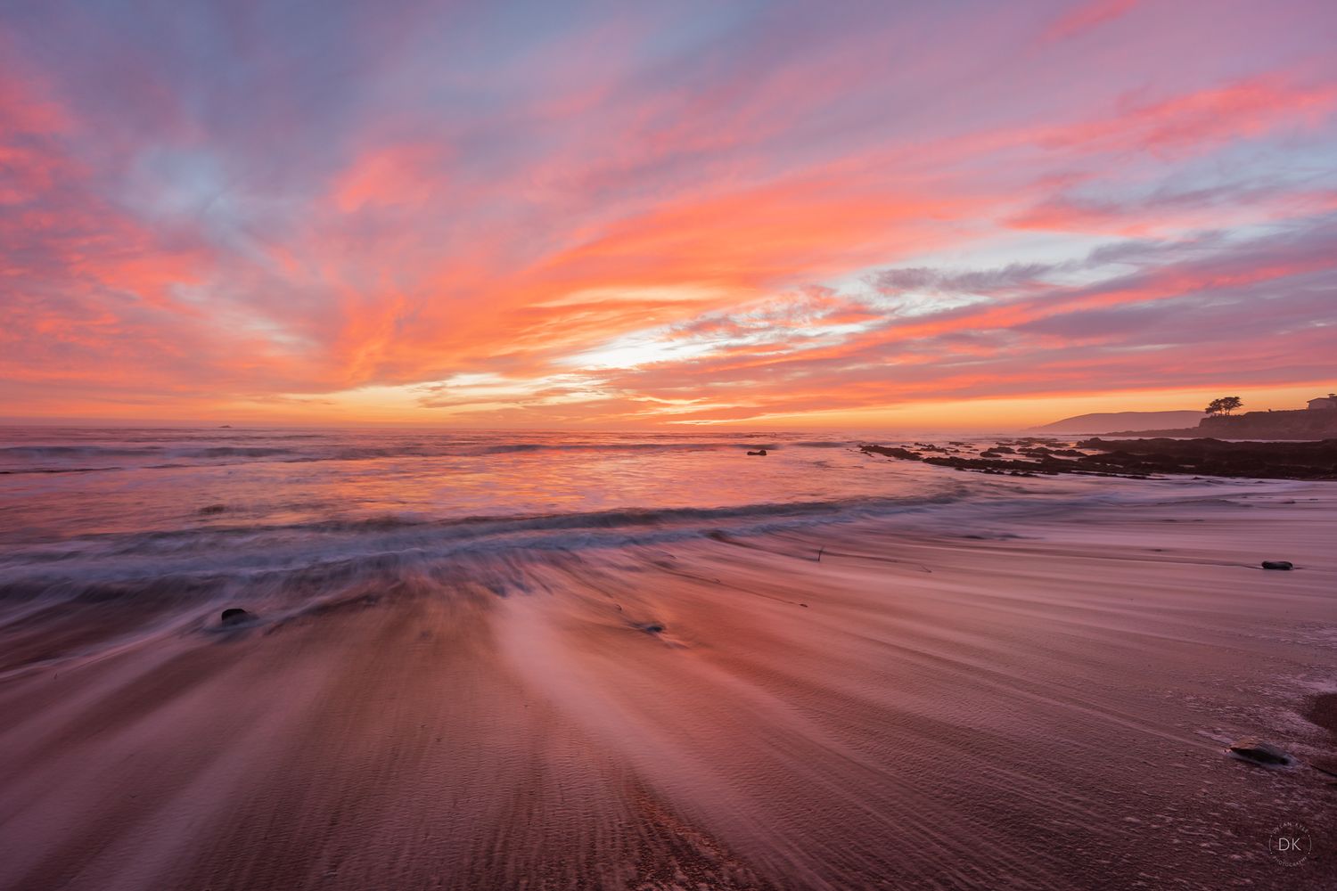 Vibrant orange and pink sunset sky reflects on wet sandy beach with waves creating dynamic patterns along the shore.