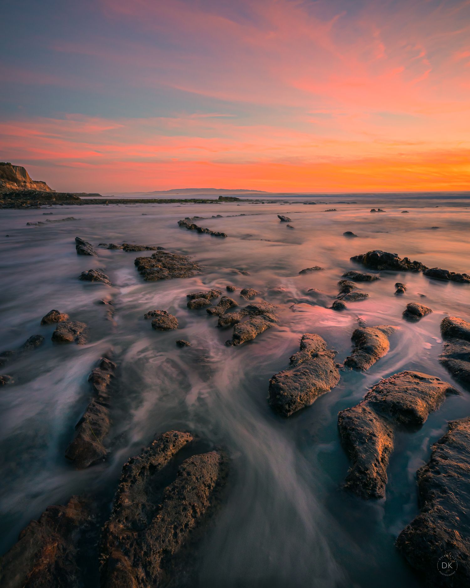 Long exposure seascape with rocky shore at sunset, capturing dramatic orange sky and smooth flowing water around rocks.