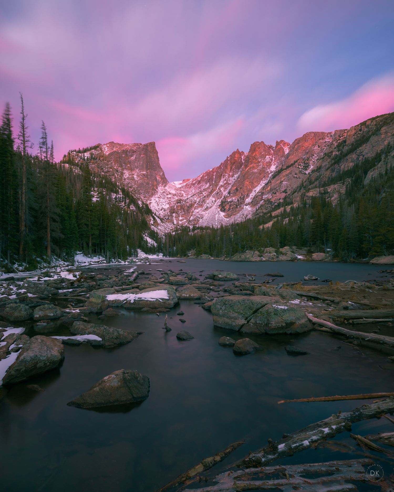 Snowy mountains reflect in calm lake waters at sunset with pink skies and scattered rocks at Rocky Mountain National Park.