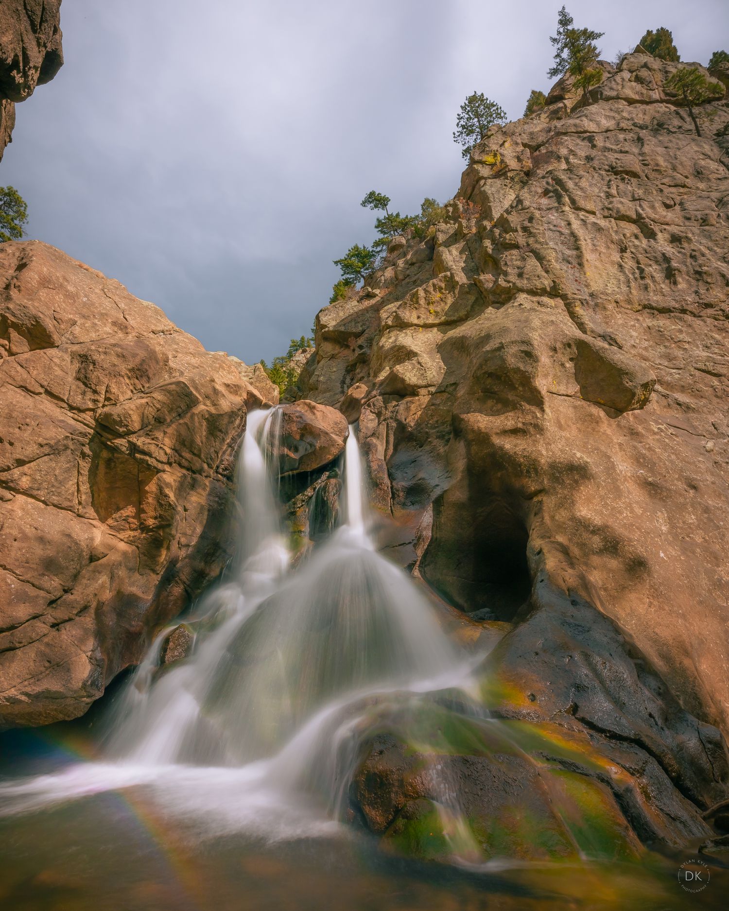 A cascading waterfall flows over orange canyon rocks with a rainbow visible in the mist at Boulder Falls, Colorado.