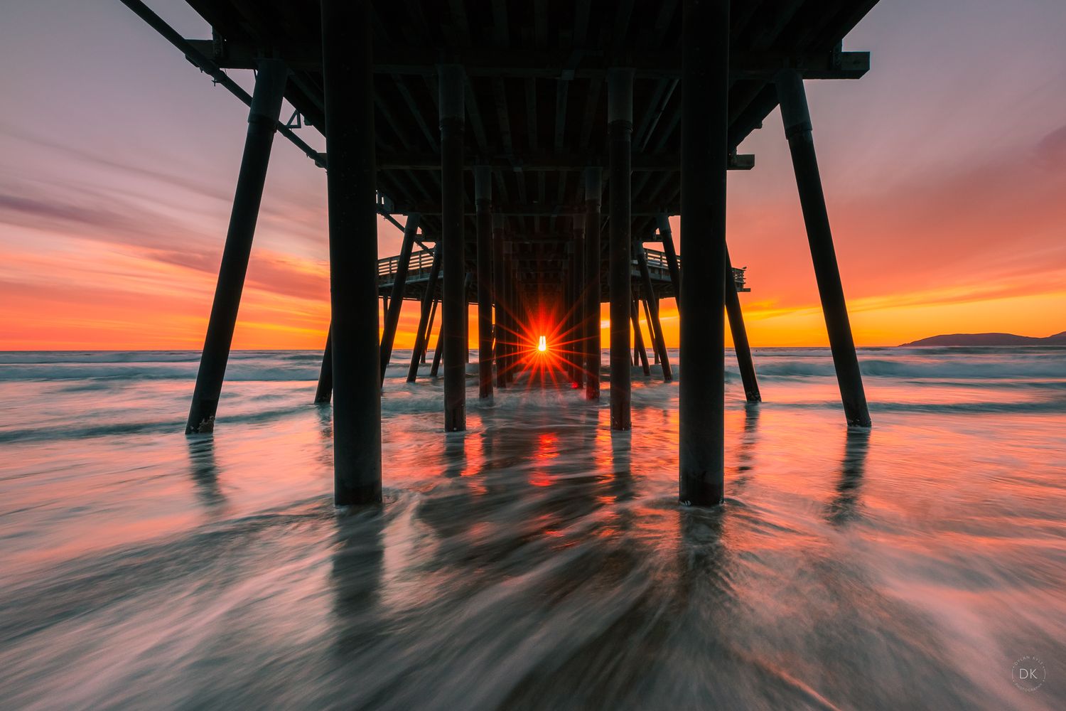 Dramatic sunset view under pier supports with silhouette against orange sky and blurred ocean waves at low tide.
