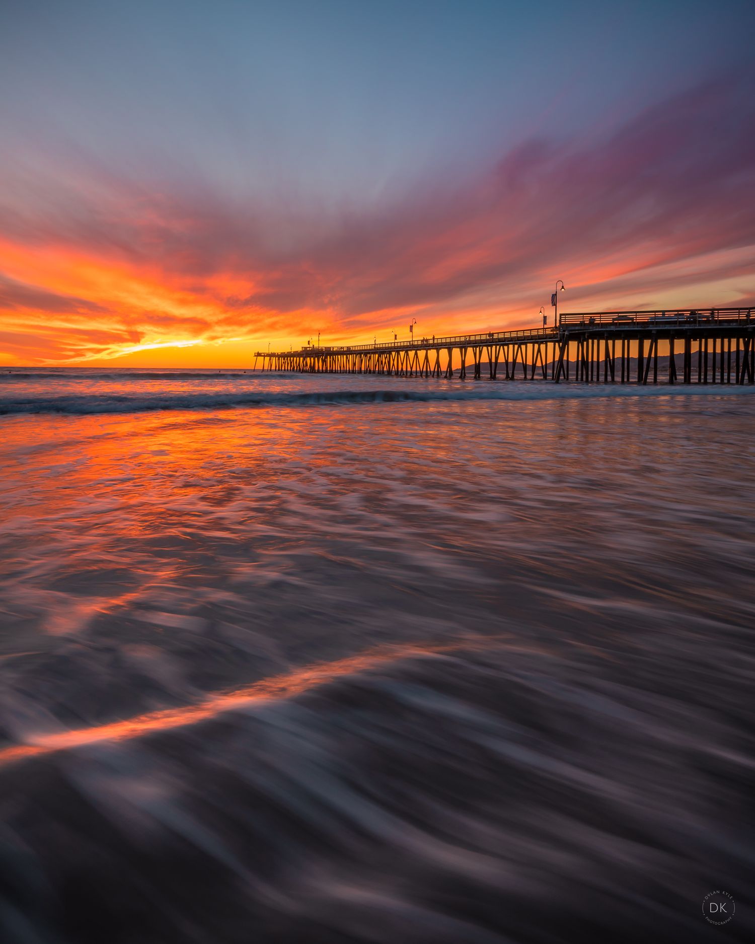Dramatic orange sunset over ocean waves with silhouette of wooden pier extending into distance at California beach.