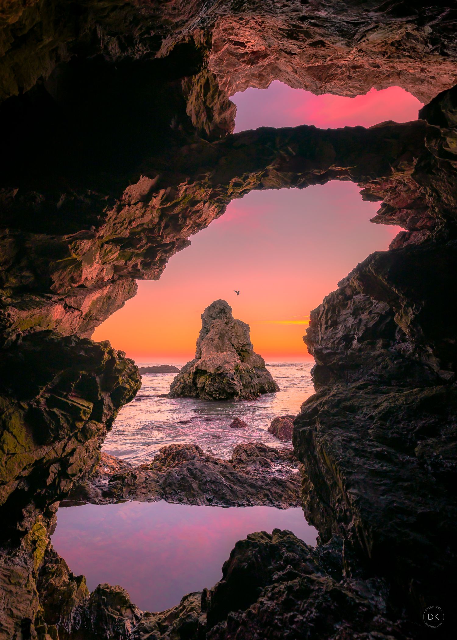 Dramatic sunset view through a sea cave opening overlooking a rocky coastline with pink and orange sky reflected in water.