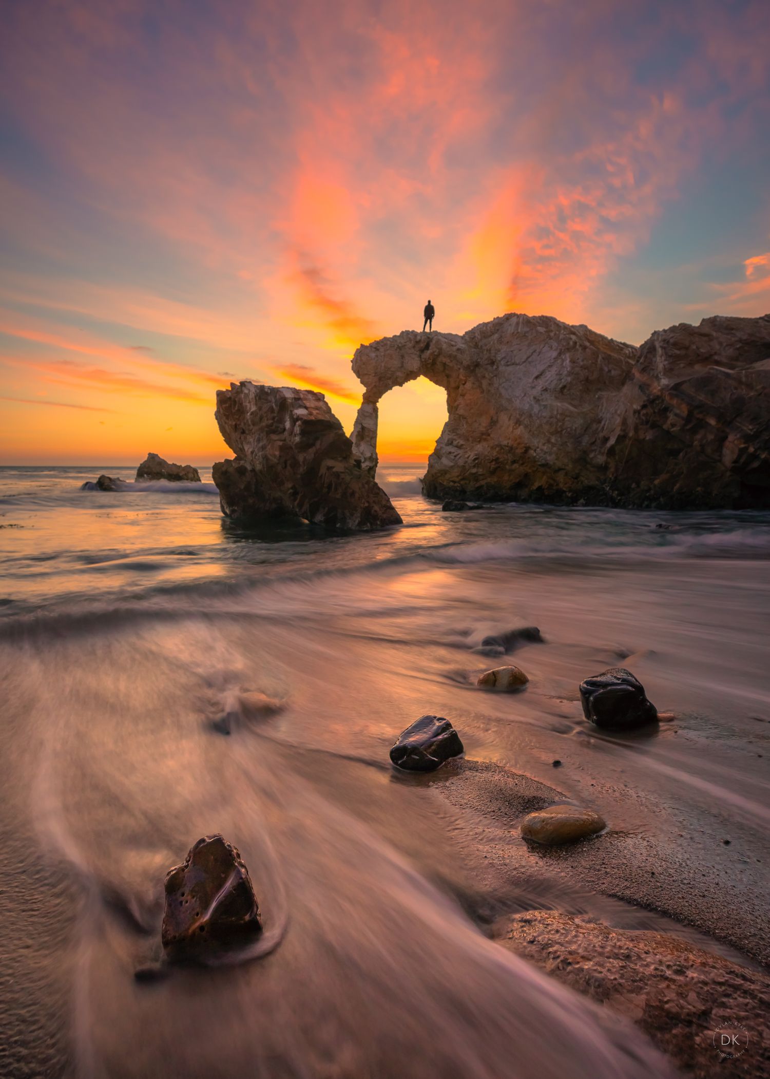 A silhouetted figure stands atop a natural rock arch over the ocean during a vibrant orange sunset at the beach.
