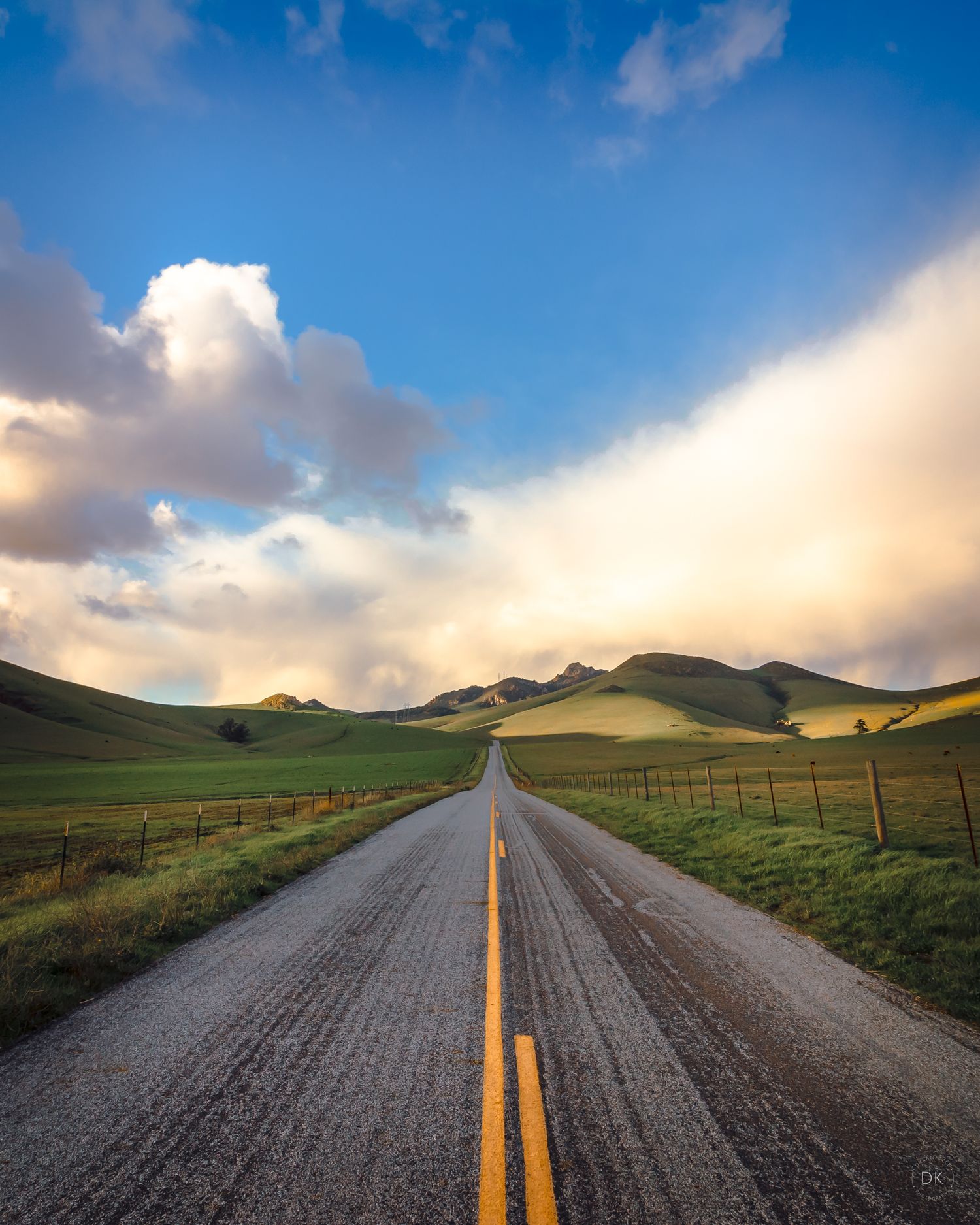 A winding rural road stretches through rolling green hills under dramatic sunset clouds and blue sky in the countryside.