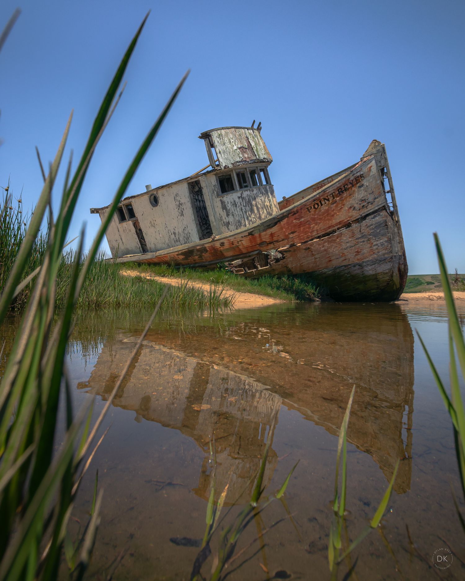 Abandoned fishing boat tilted on muddy shore with marsh grass and reflections in calm water at sunset.