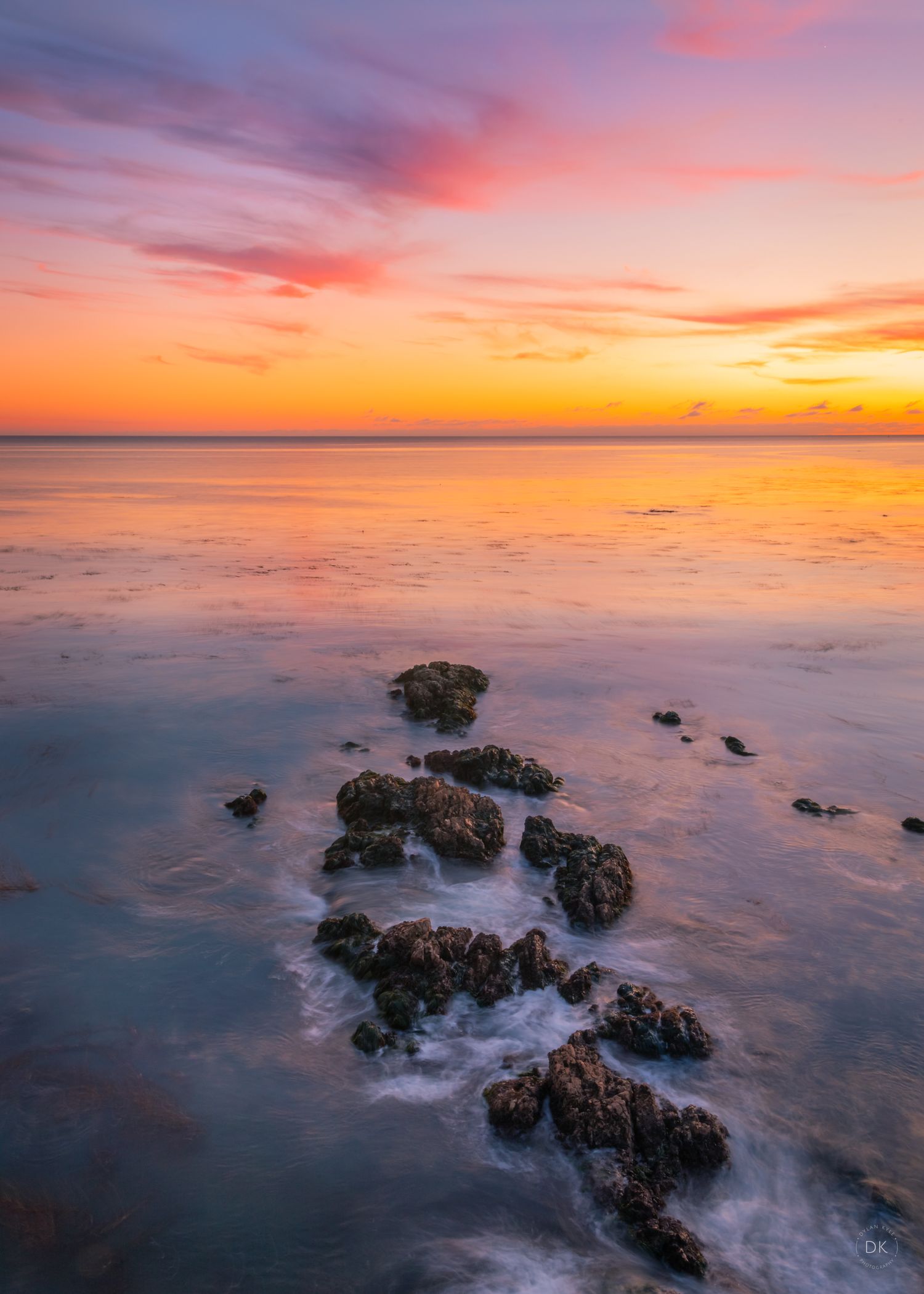 Dramatic sunset over calm ocean waters with rocky outcrops creating gentle waves in the foreground.