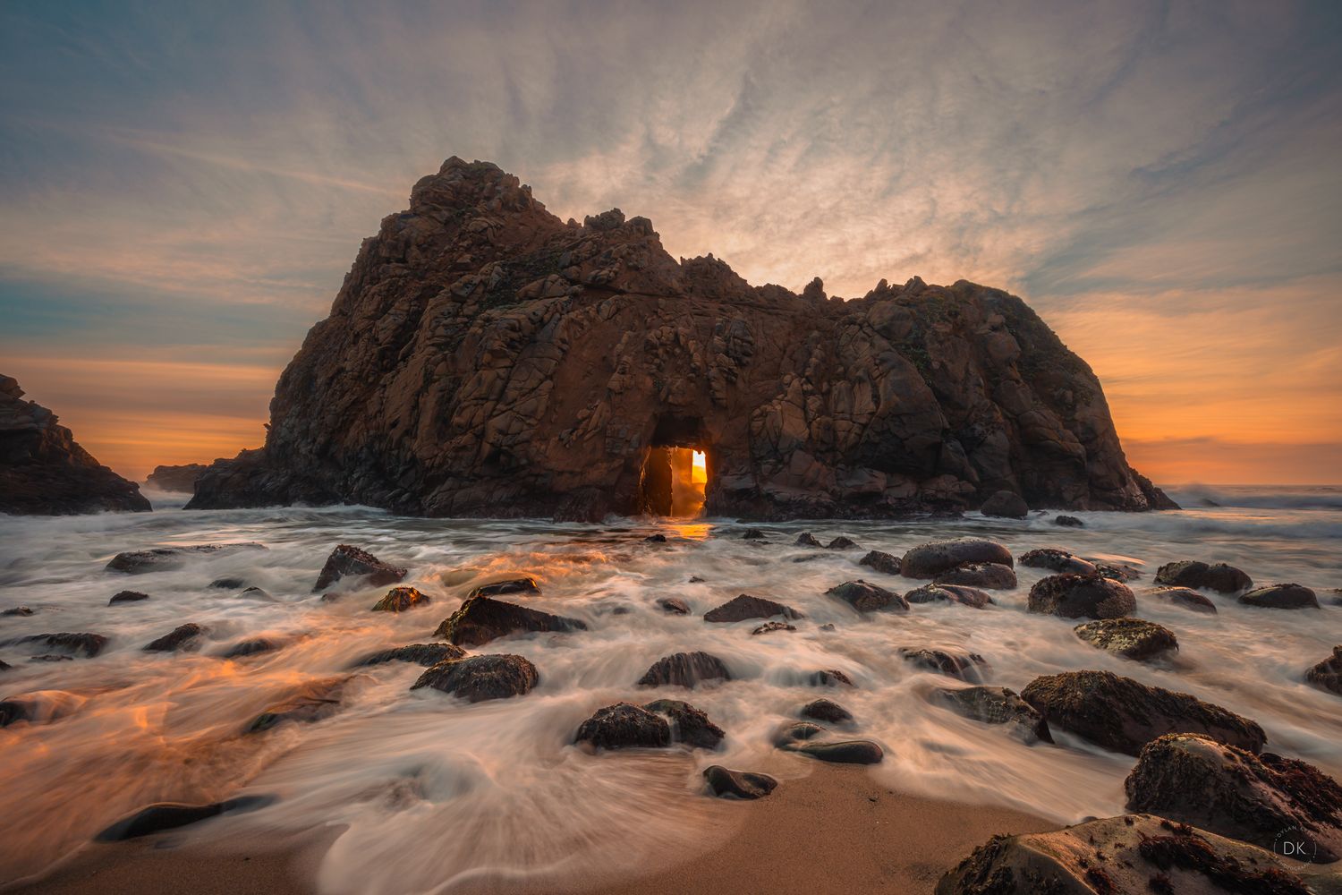 Sunset light streams through natural rock arch at Pfeiffer Beach with waves flowing over smooth stones in Big Sur, California.